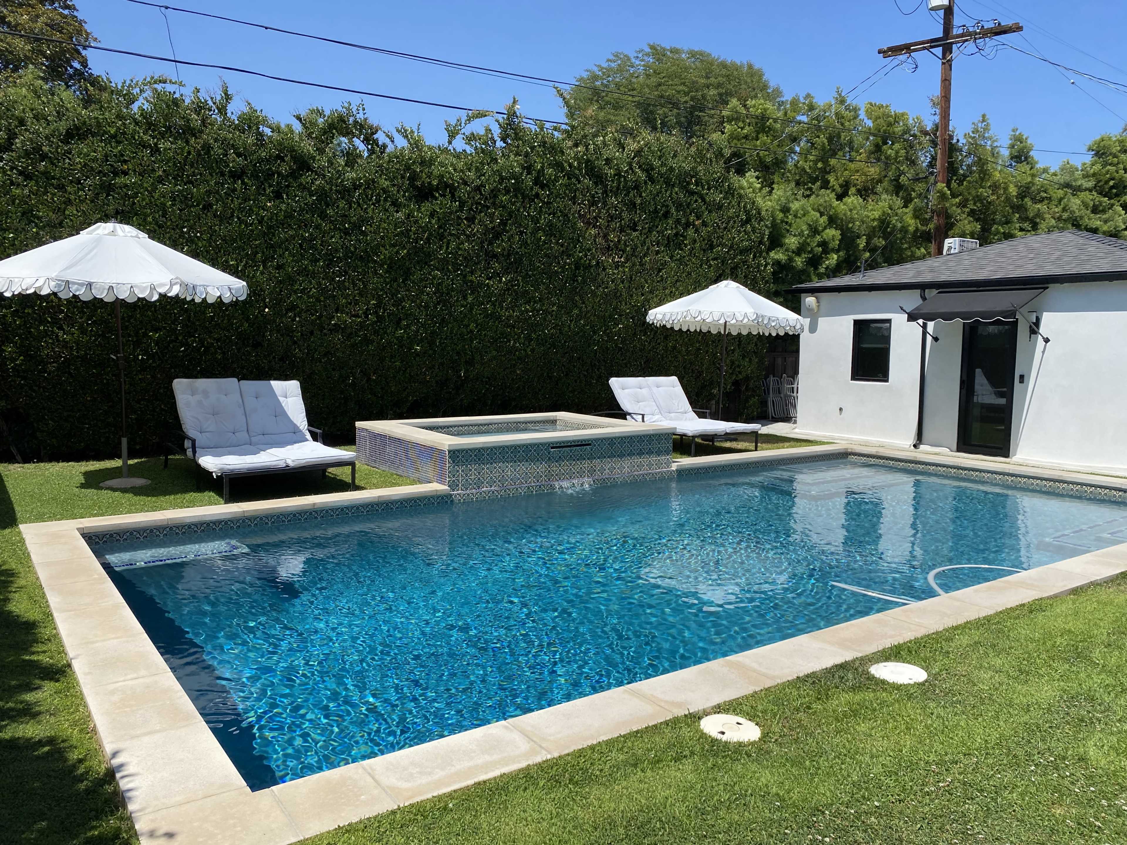 A swimming pool surrounded by lounge chairs and two umbrellas, with a small spa area and a nearby structure set against a backdrop of greenery.