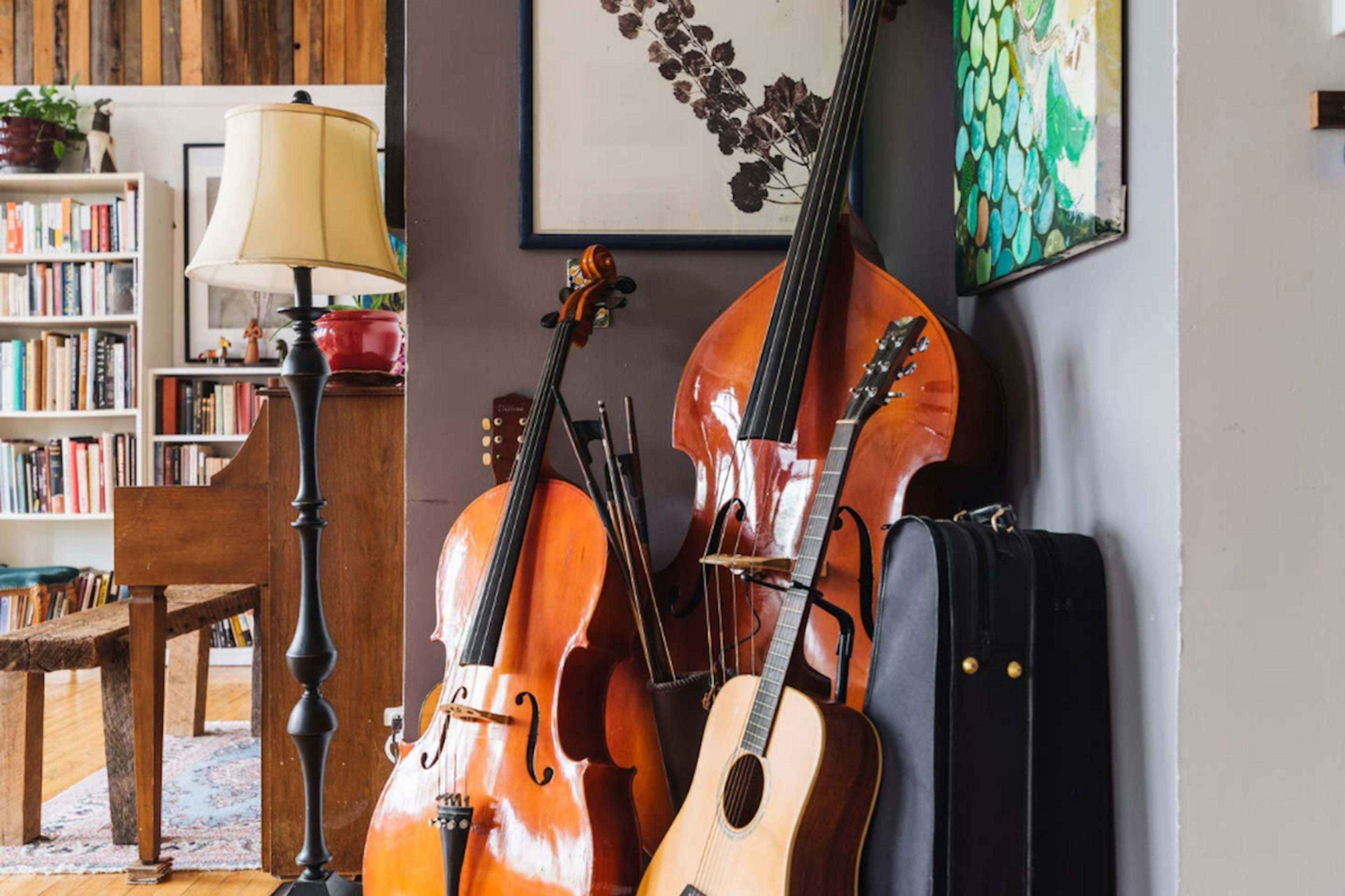 The image shows a collection of stringed instruments, including a cello, double bass, and guitar, placed next to a standing lamp in a cozy interior setting.