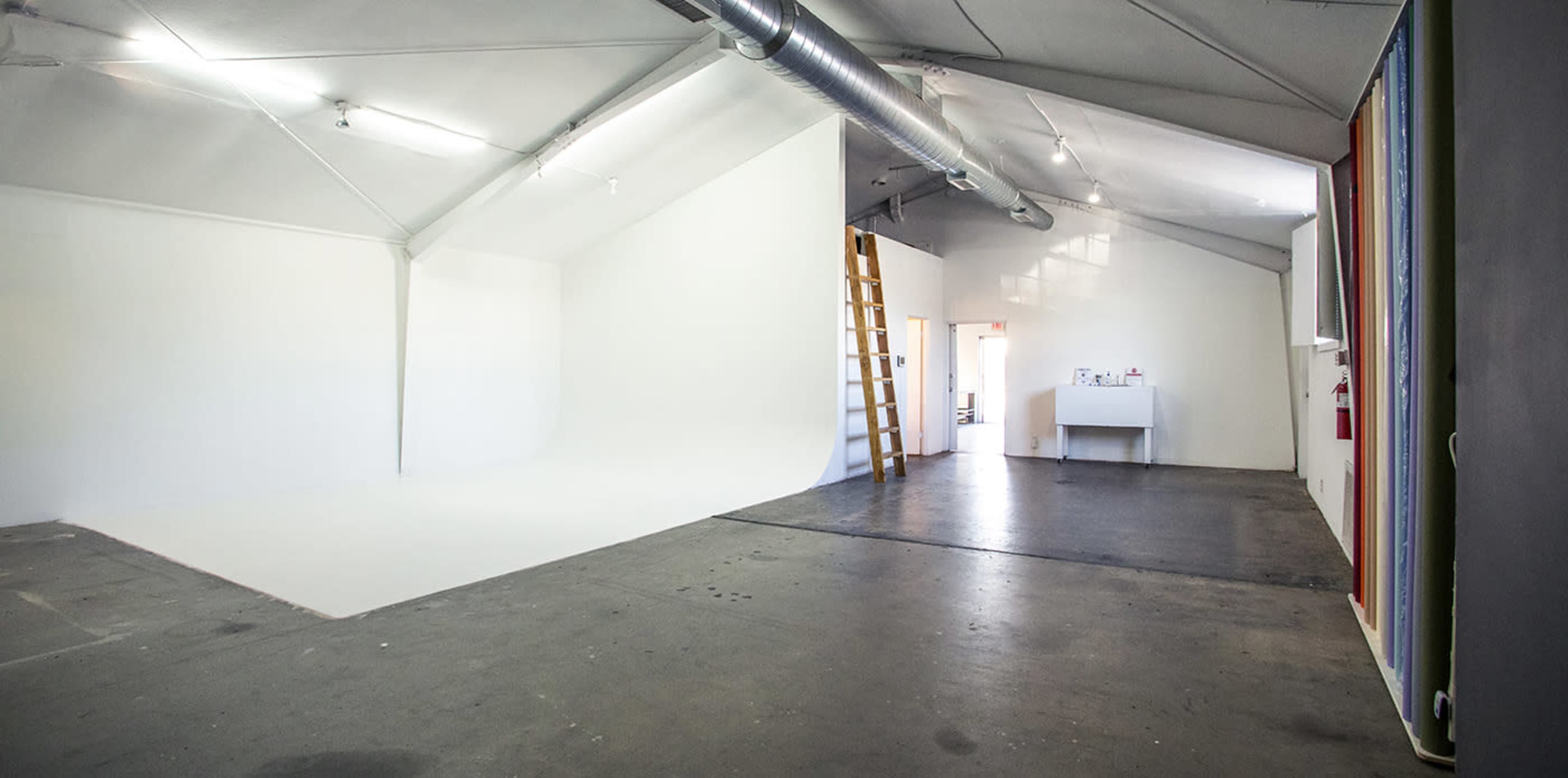 A spacious studio with a white backdrop on one side, a ladder leaning against the wall, and a small workstation in the background.