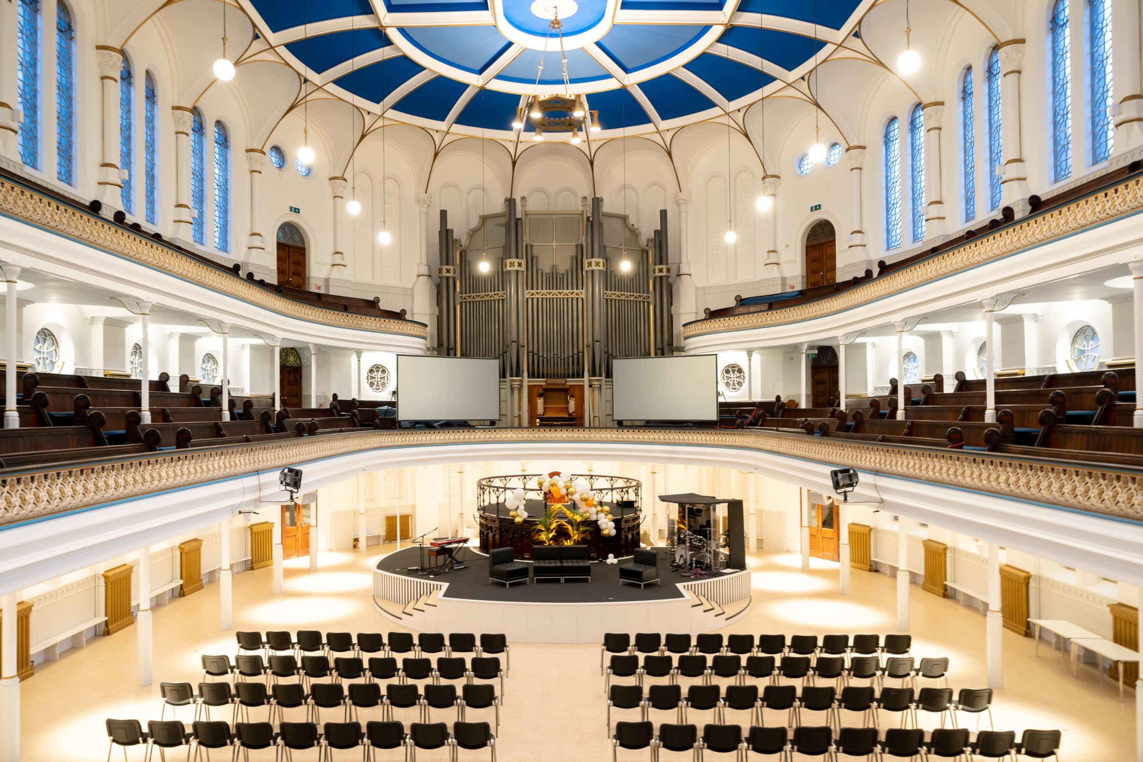 The interior of a large, well-lit auditorium with a stage at the front, surrounded by rows of black chairs and an organ in the background.