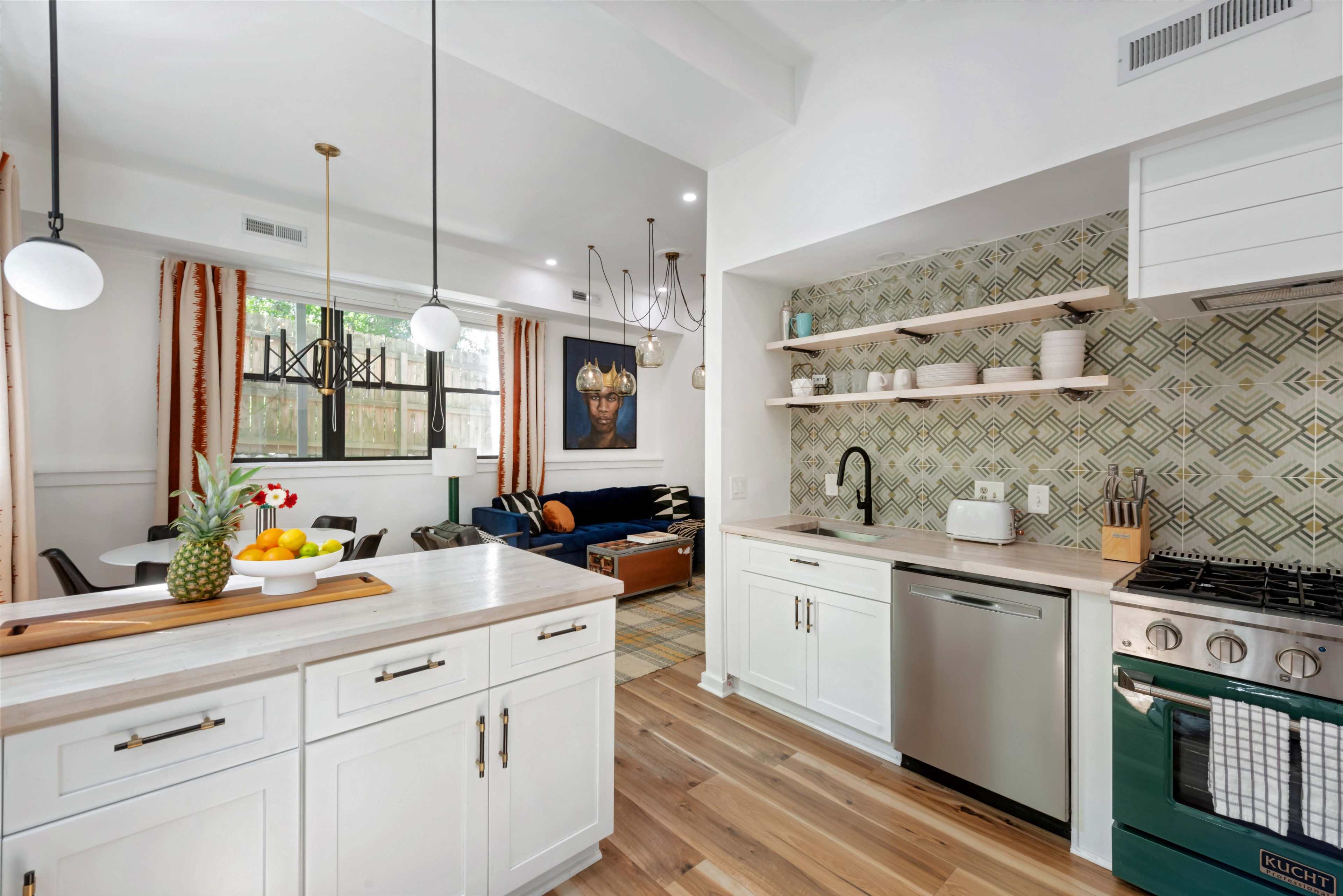 The image features a modern kitchen with white cabinetry and a green stove, seamlessly connected to a living area that includes a dark sofa and decorative artwork on the wall.