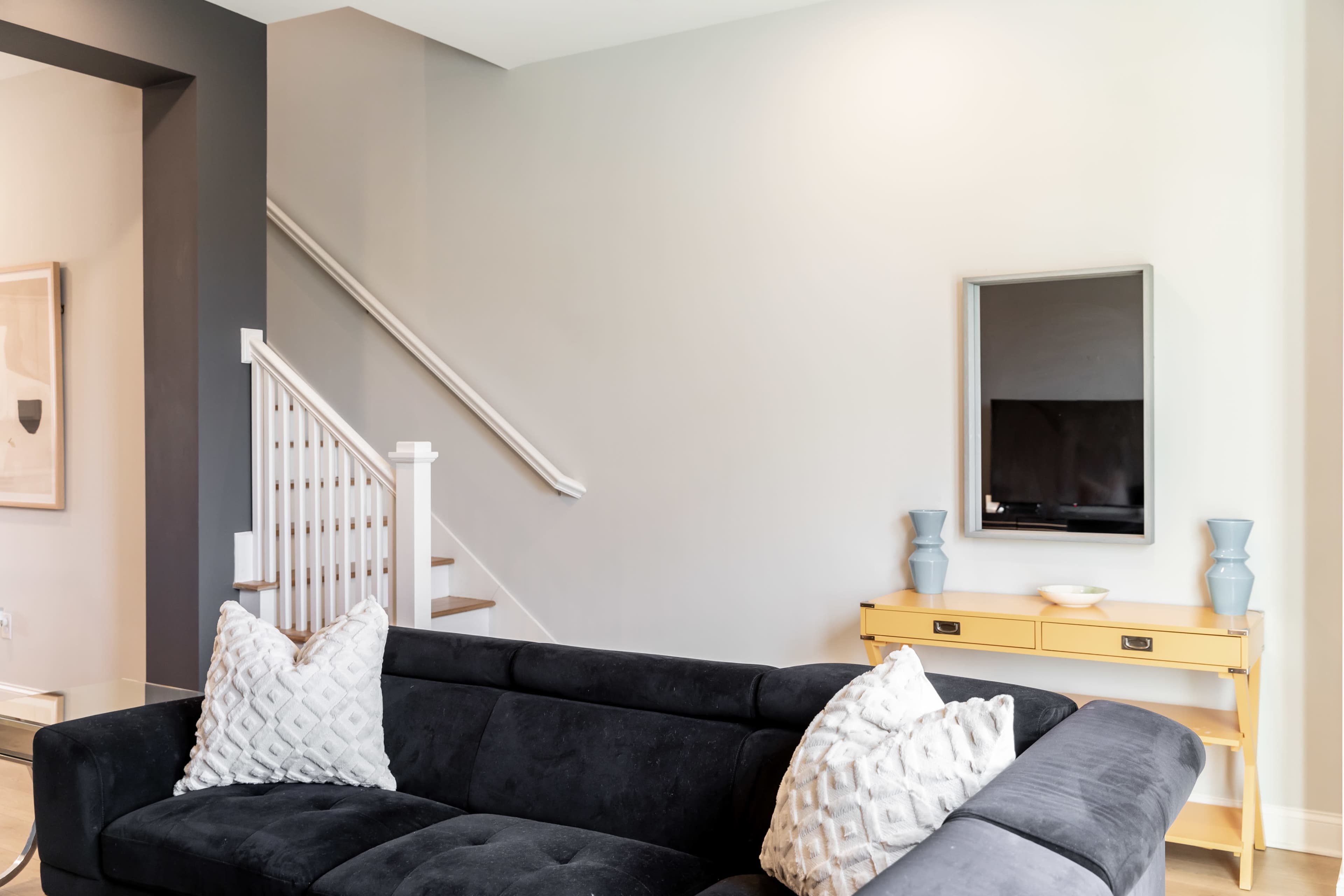 A dark sectional sofa with decorative pillows faces a yellow side table and a television mounted on a light gray wall, with a staircase visible in the background.