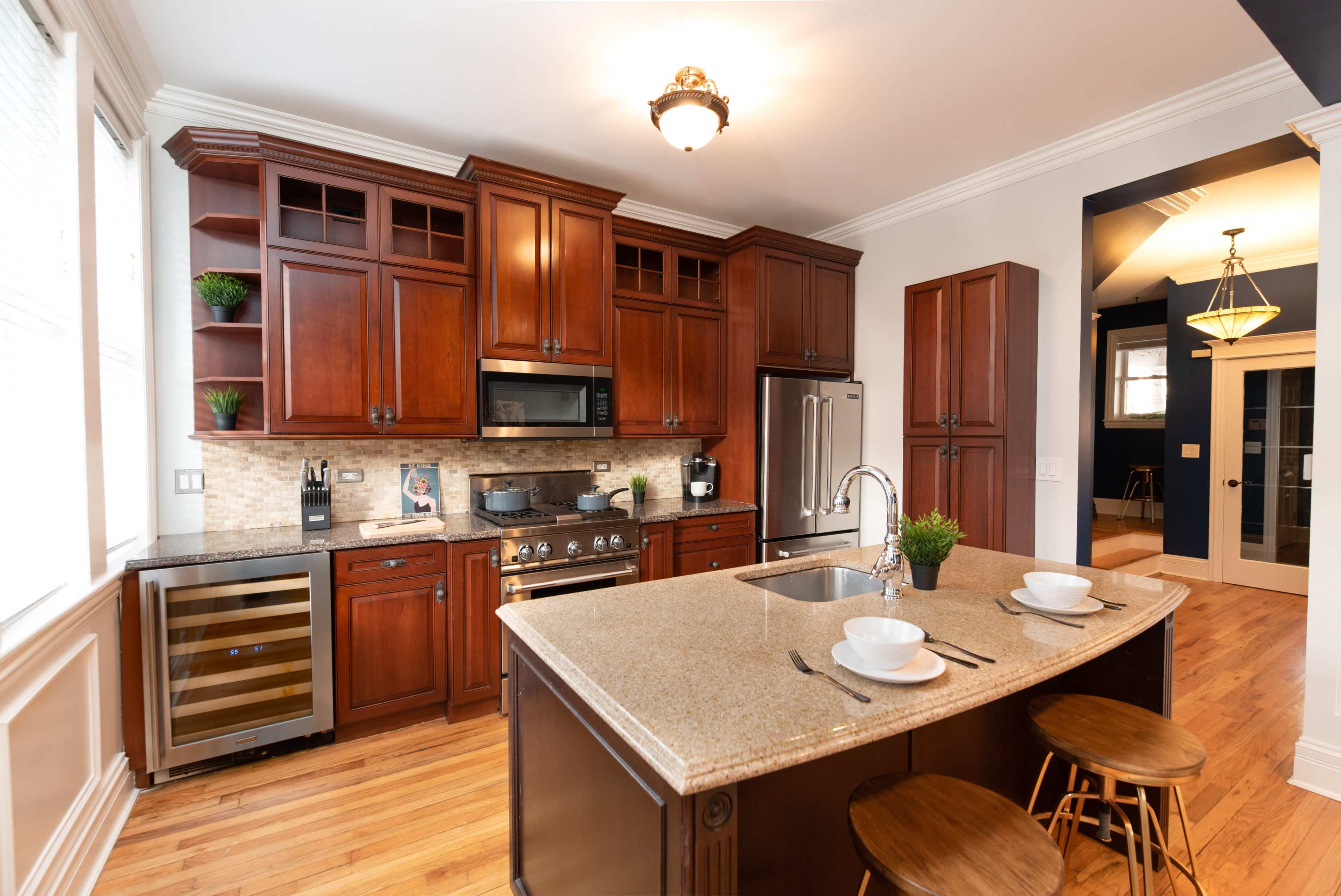 The image shows a modern kitchen featuring dark wooden cabinetry, a large island with a granite countertop, bar stools, and stainless steel appliances.