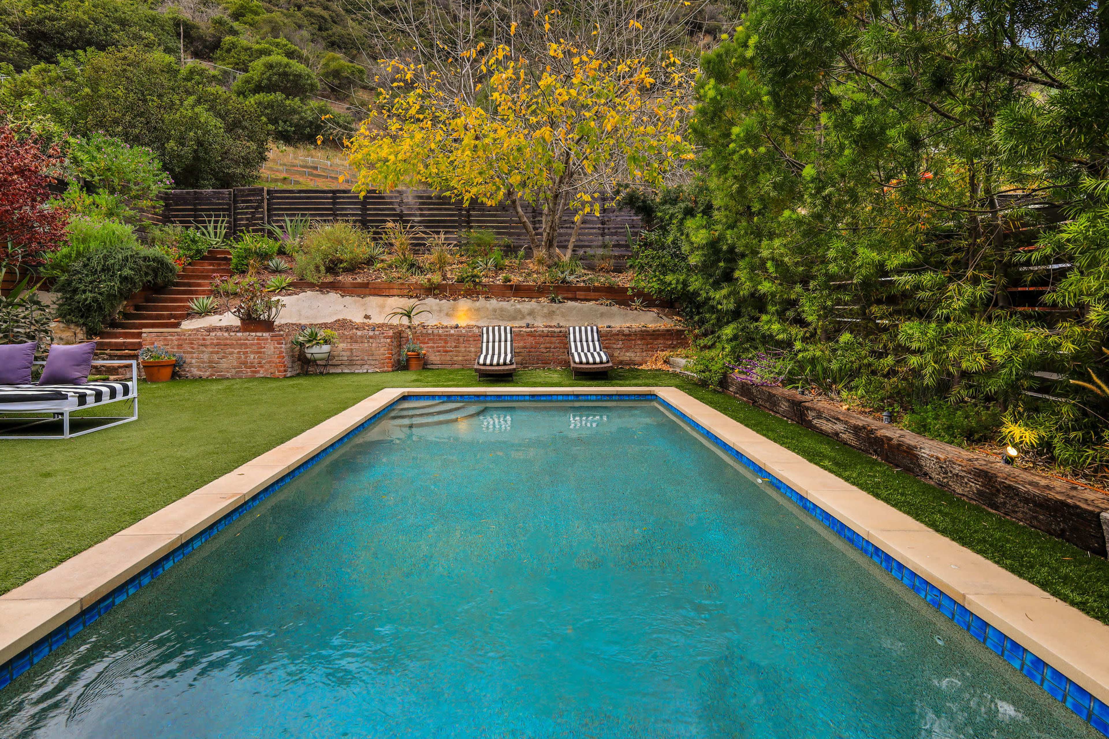 A rectangular swimming pool is set in a landscaped yard featuring green grass, lounge chairs, and a backdrop of trees and garden beds.
