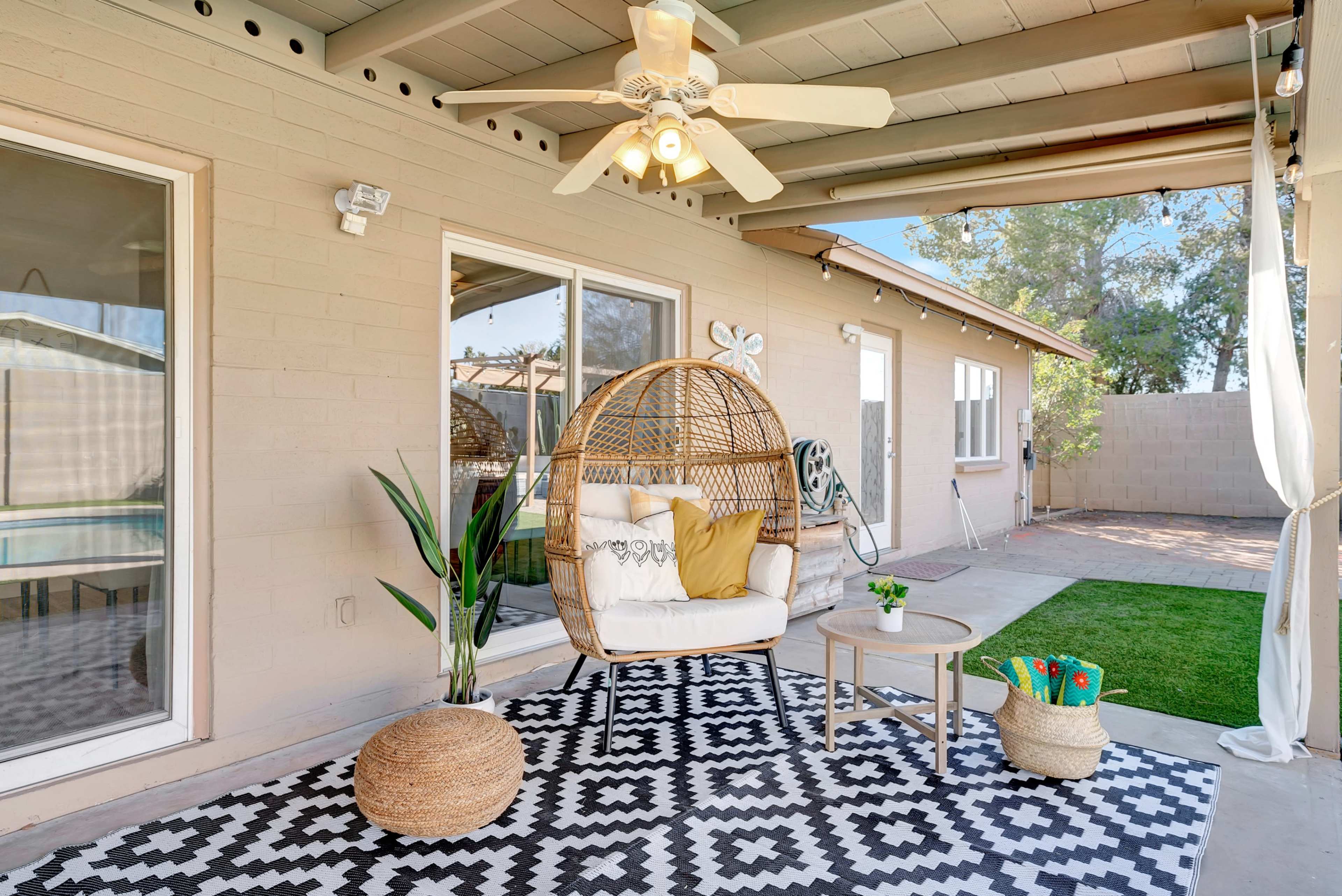 A covered patio features a hanging rattan chair with cushions, a small table, and potted plants, set on a geometric-patterned rug.