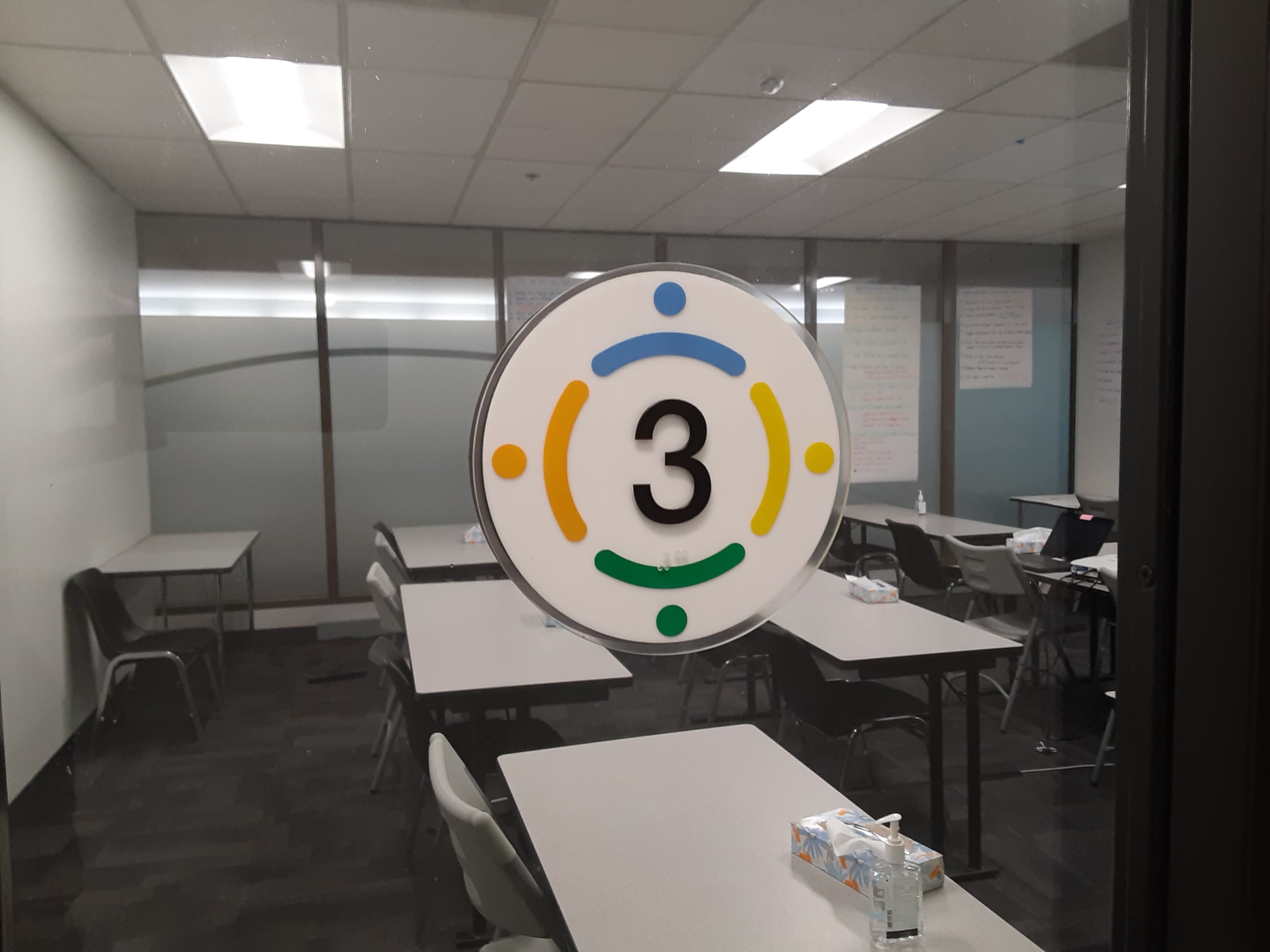 The image shows a circular sign with the number "3" and colored dots in blue, yellow, and green, affixed to a glass door leading to a classroom or meeting space with tables and chairs visible inside.
