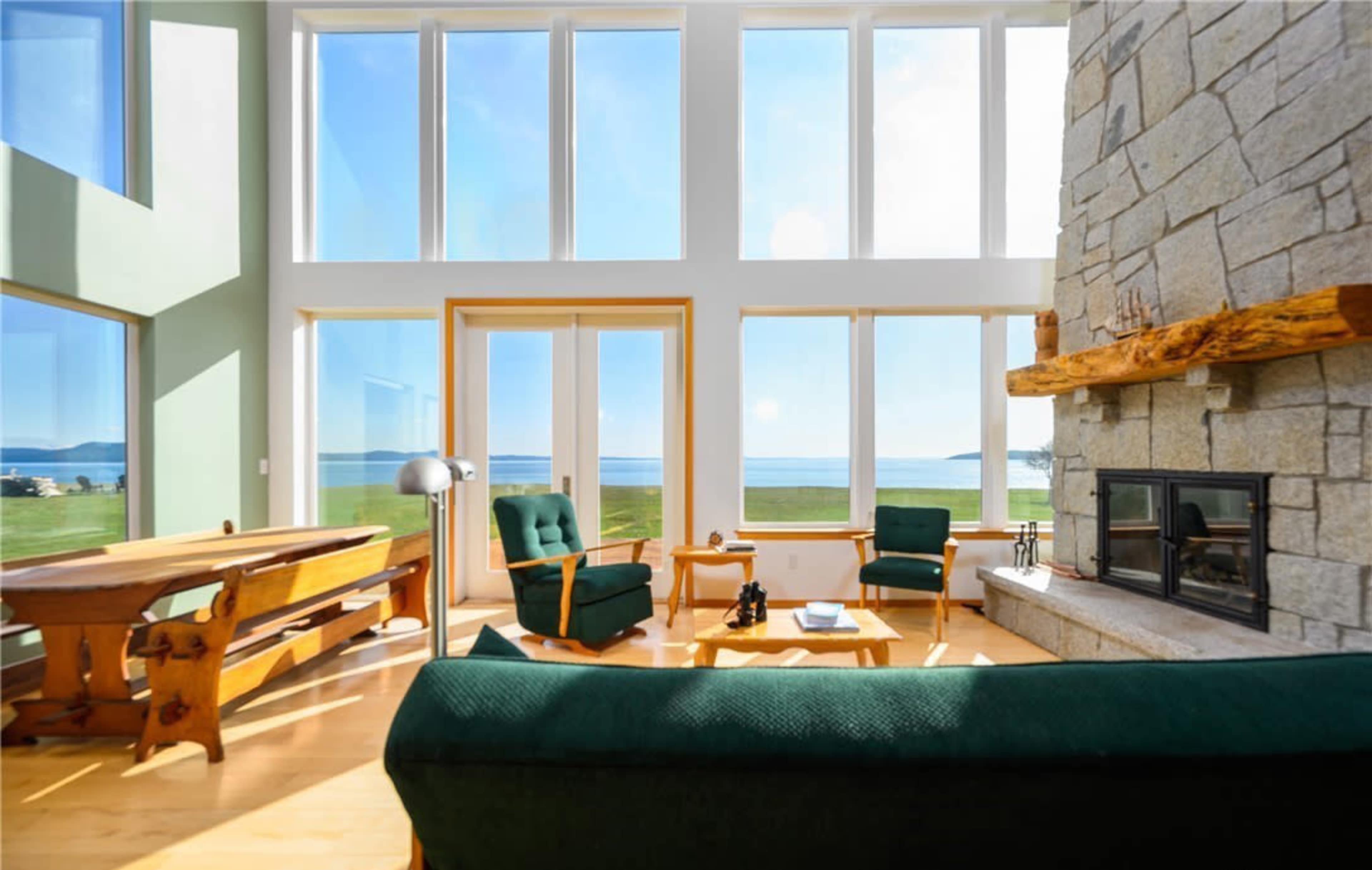 A spacious living room features large windows overlooking a scenic view, a stone fireplace, a wooden table, and green chairs.