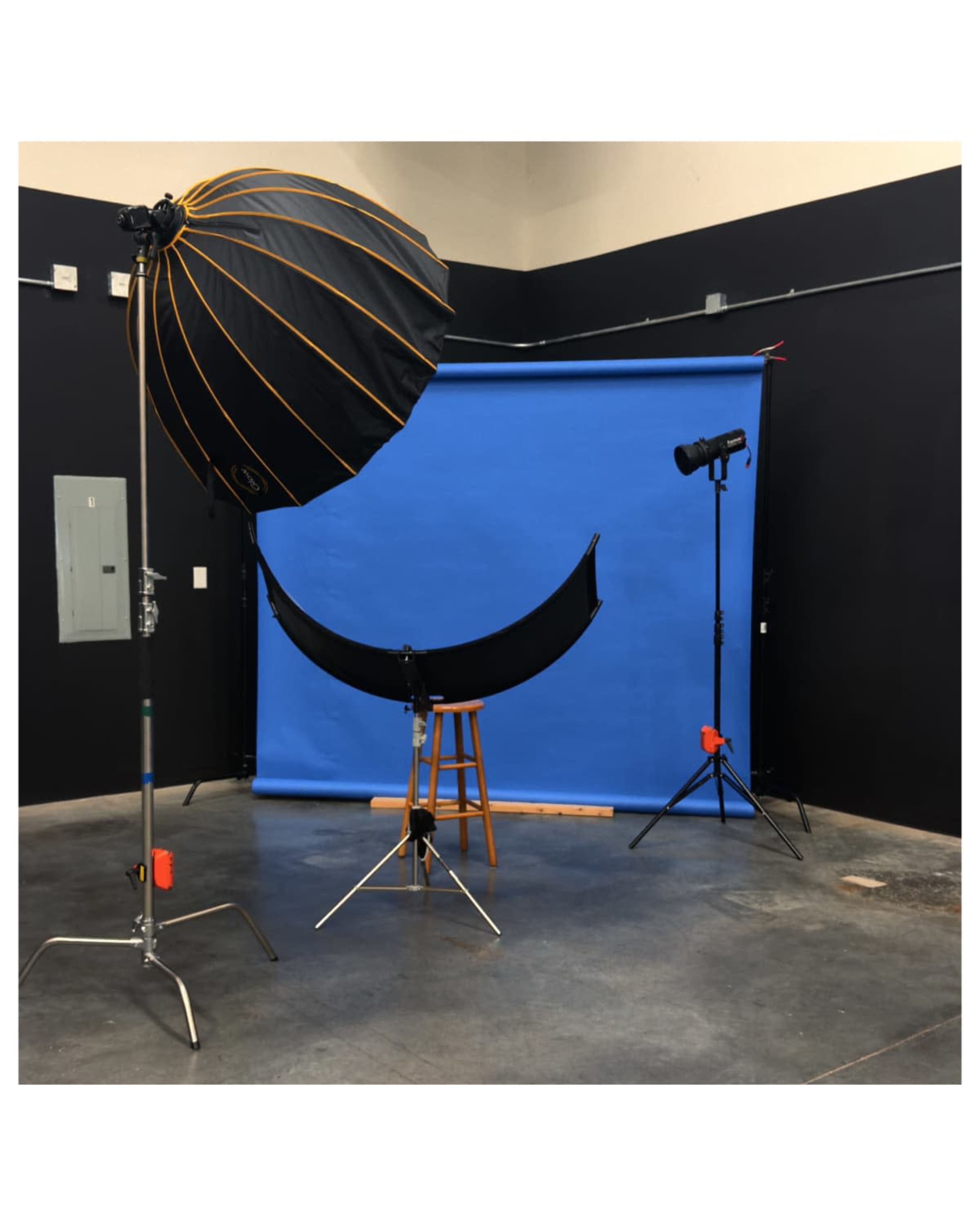 The image shows a photography studio setup with a blue backdrop, a black umbrella light, a stool, and a secondary light on a stand.