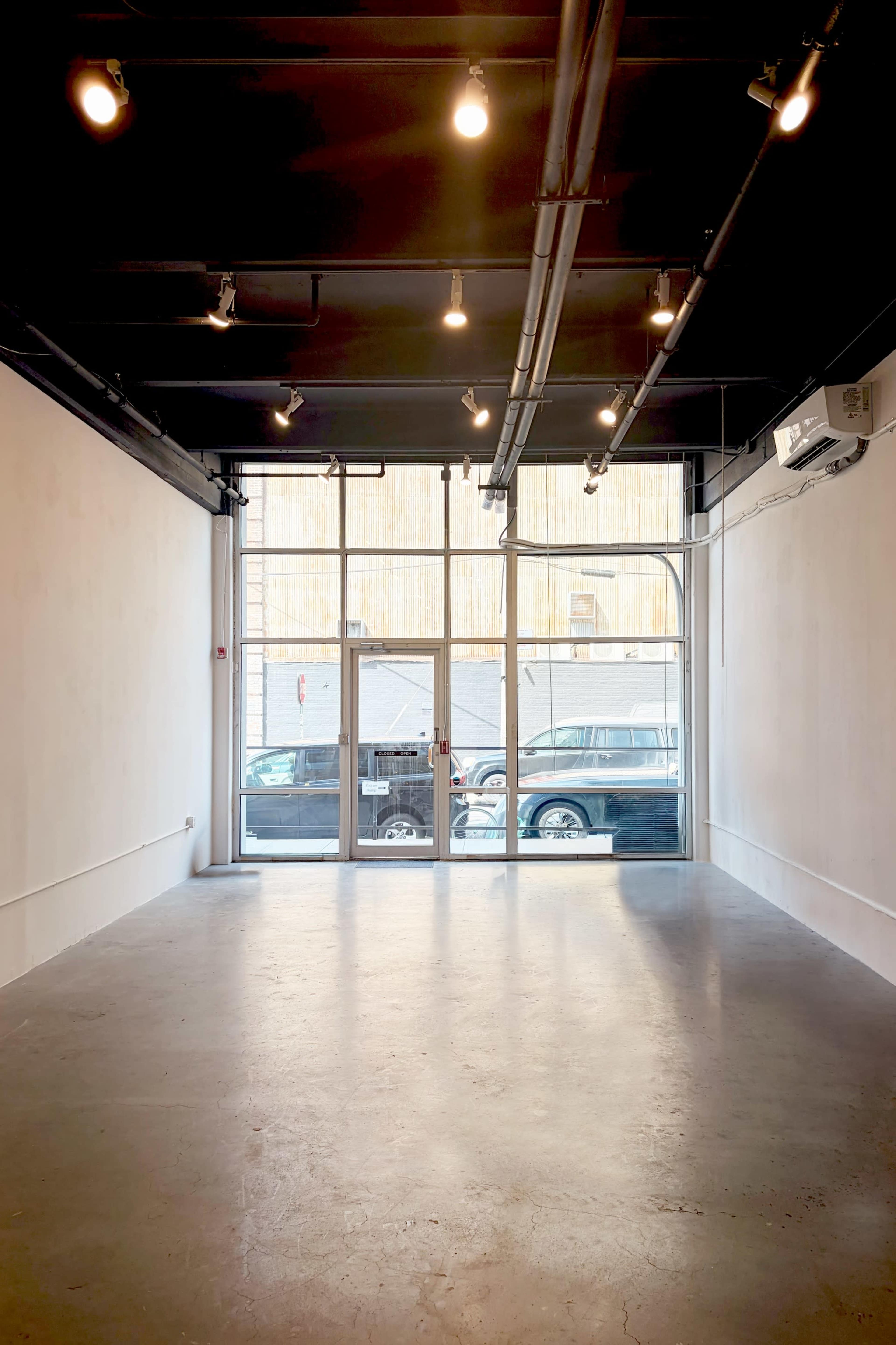 The image shows a vacant commercial space with concrete flooring, large front windows, and overhead lights.