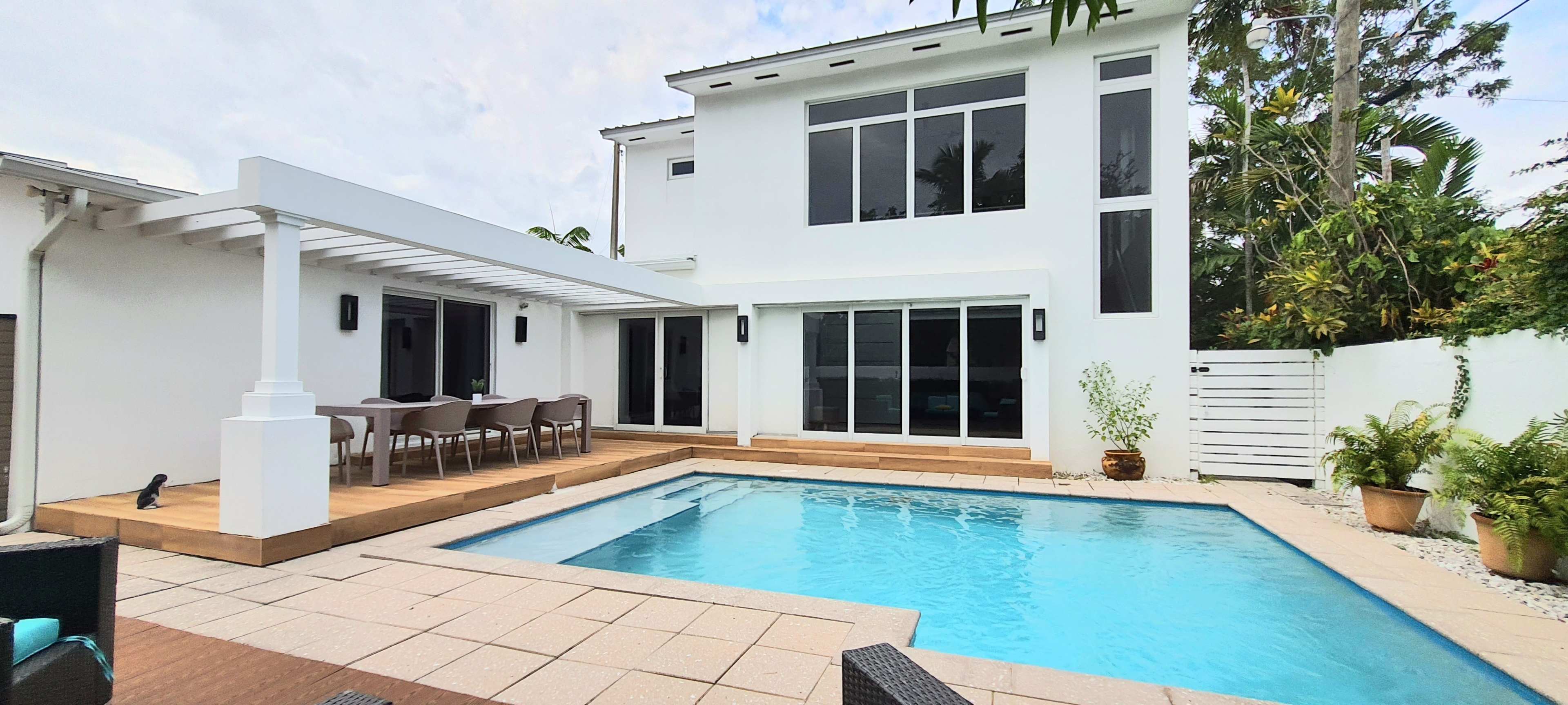 The image shows a modern home with a swimming pool, outdoor seating area, and large windows surrounded by tropical plants.