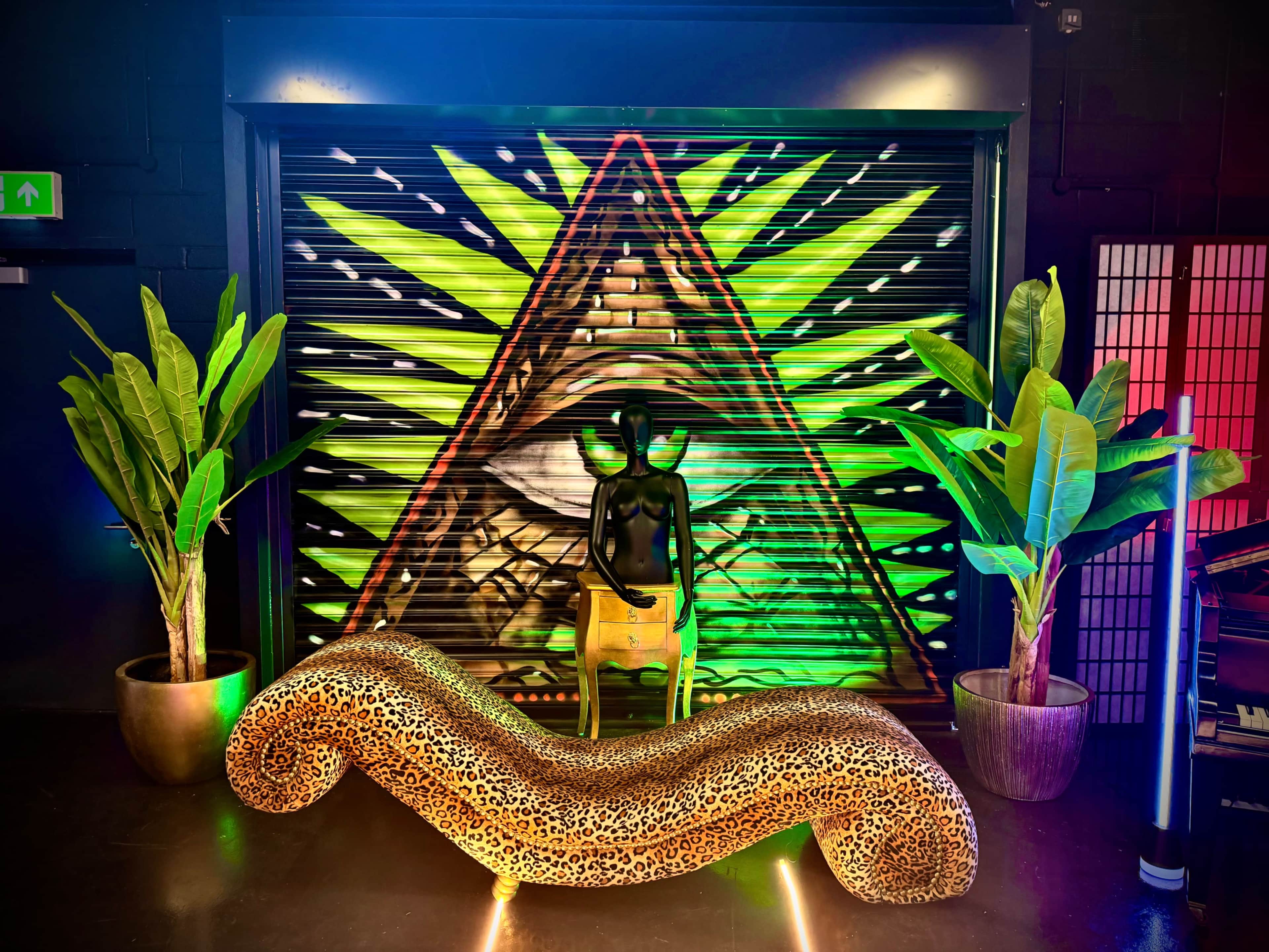 The image features a black mannequin standing near a leopard-print lounge chair, positioned in front of a vibrant, eye-catching mural with an eye and foliage design.