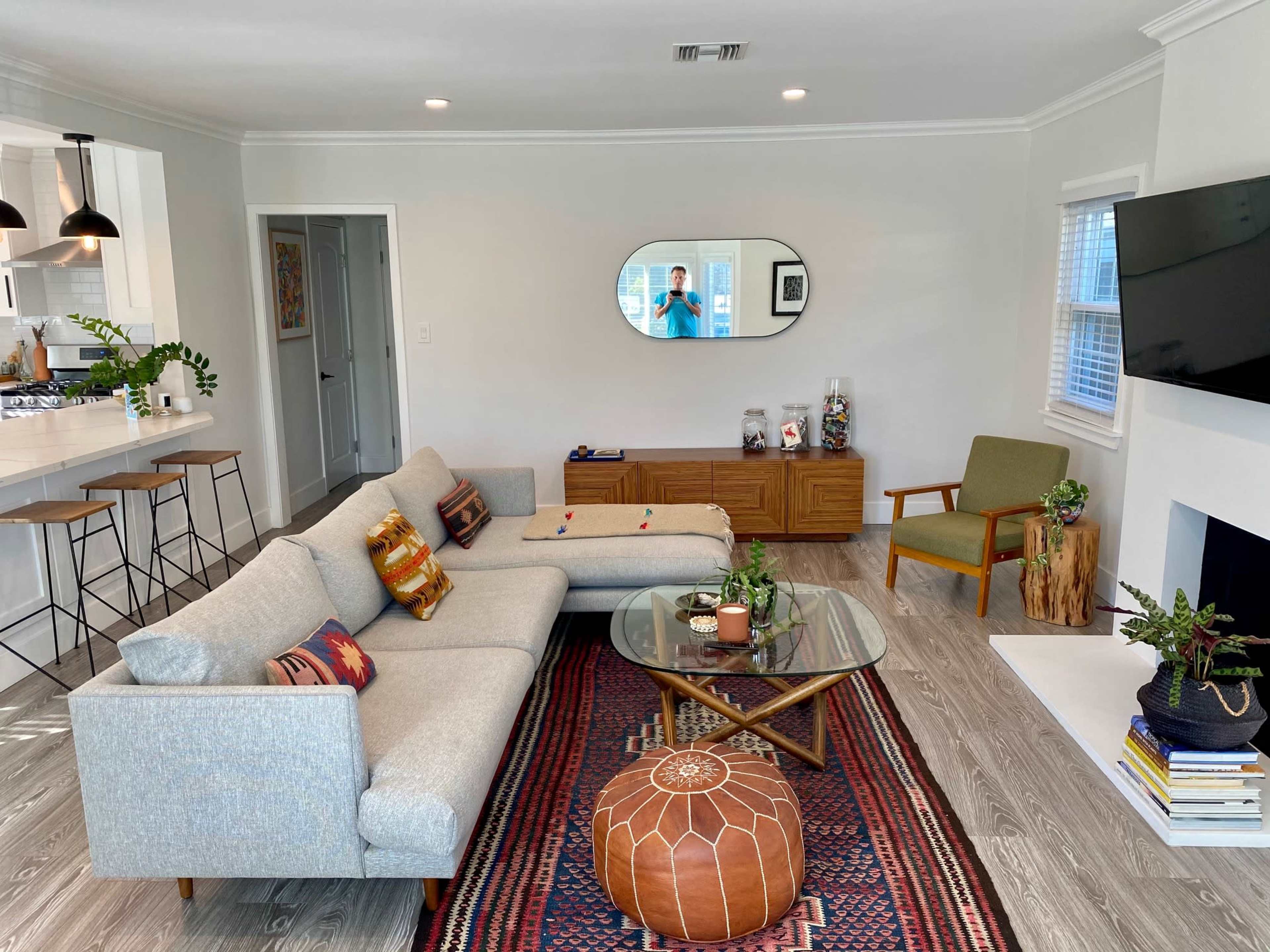 A modern living room features a light gray sectional sofa, a glass coffee table, a patterned rug, and a wall-mounted TV, with plants and decorative objects on display.