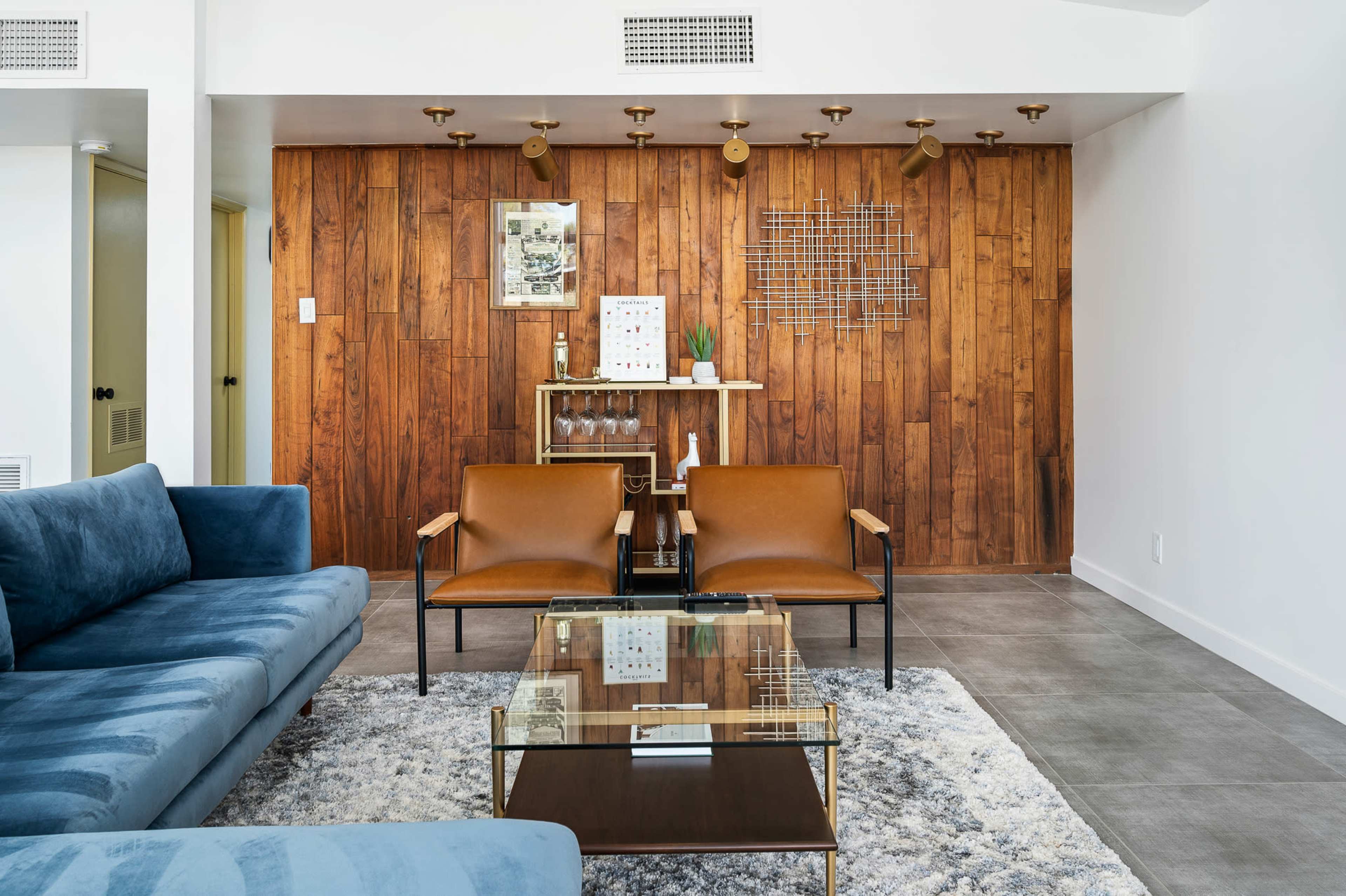 A modern living room with a blue sofa, two brown chairs, a glass coffee table, and a wooden accent wall featuring decorative shelves.