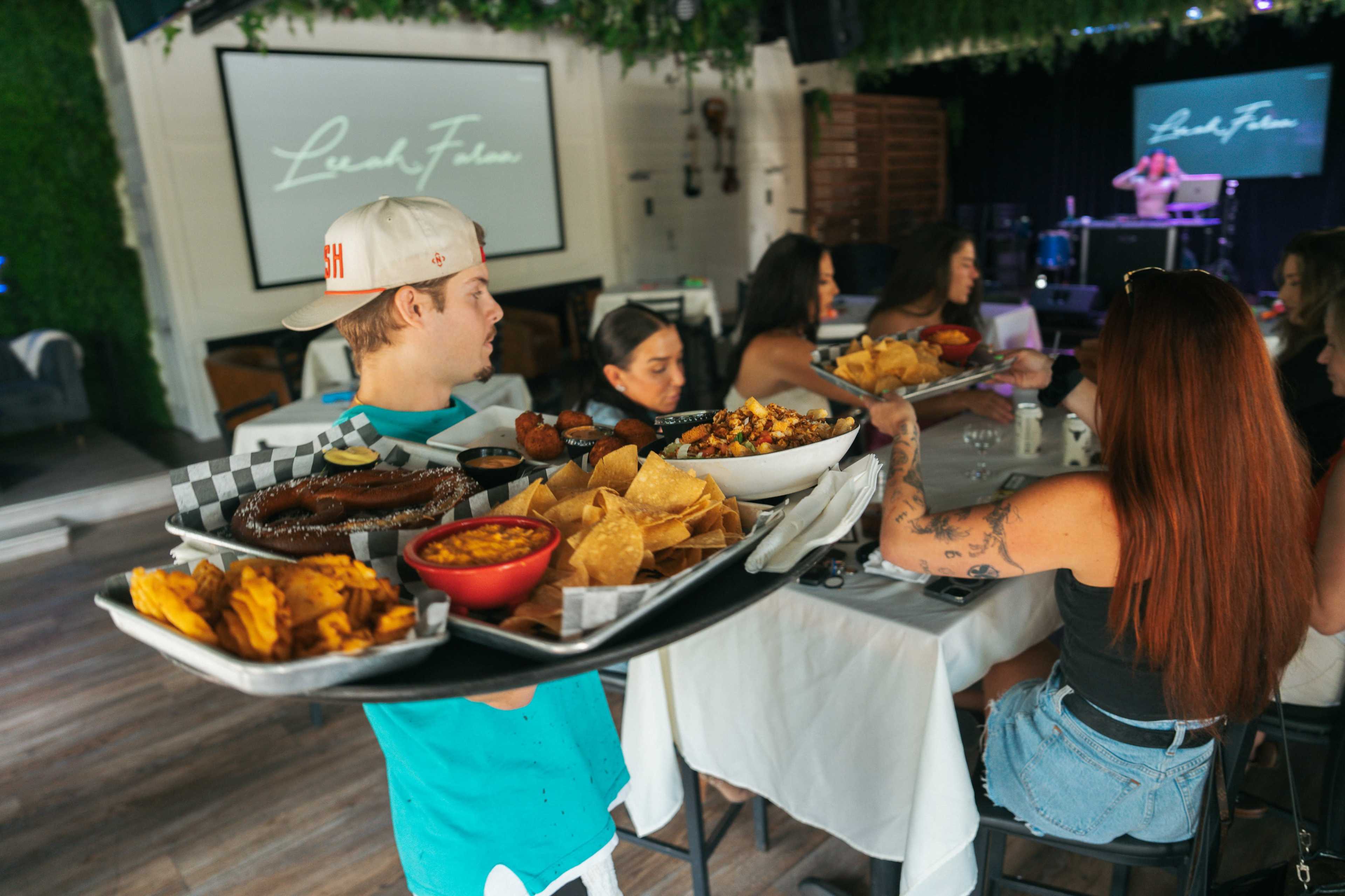 A server carries a large tray filled with various dishes, including nachos and fried foods, to a group of diners seated at a table in a restaurant.