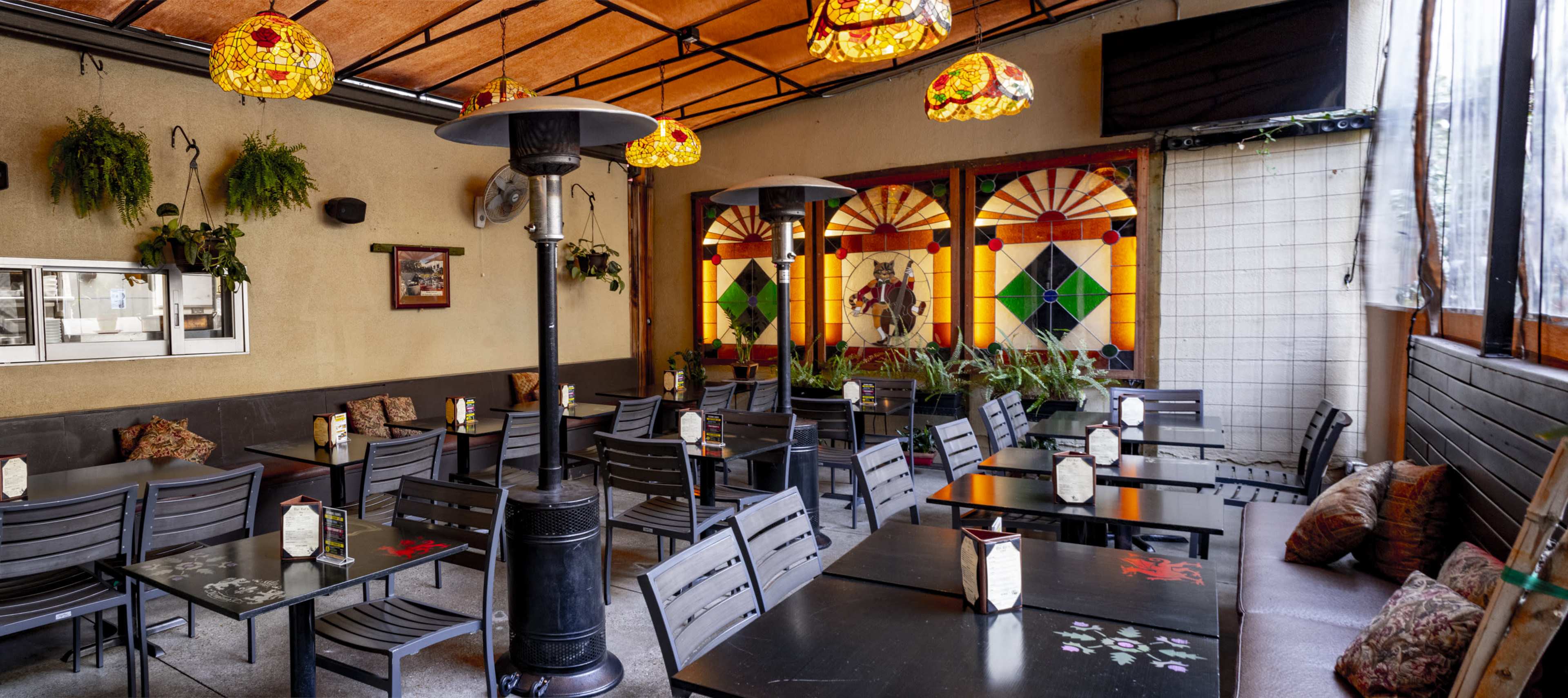 The interior of a restaurant features rows of black tables and chairs, decorative stained glass windows, and pendant lamps hanging from a wooden ceiling.