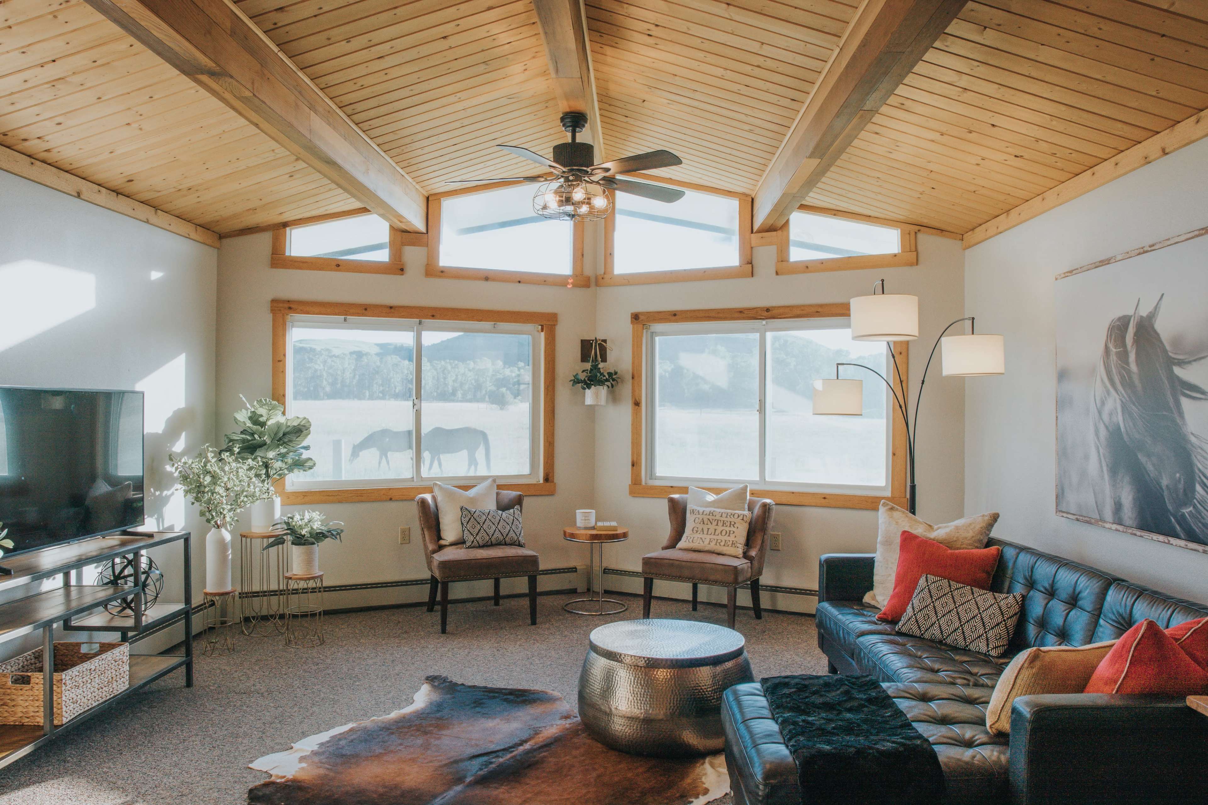 A modern living room features a leather couch, two chairs, a large window with a view of horses outside, and wooden beams on the ceiling.
