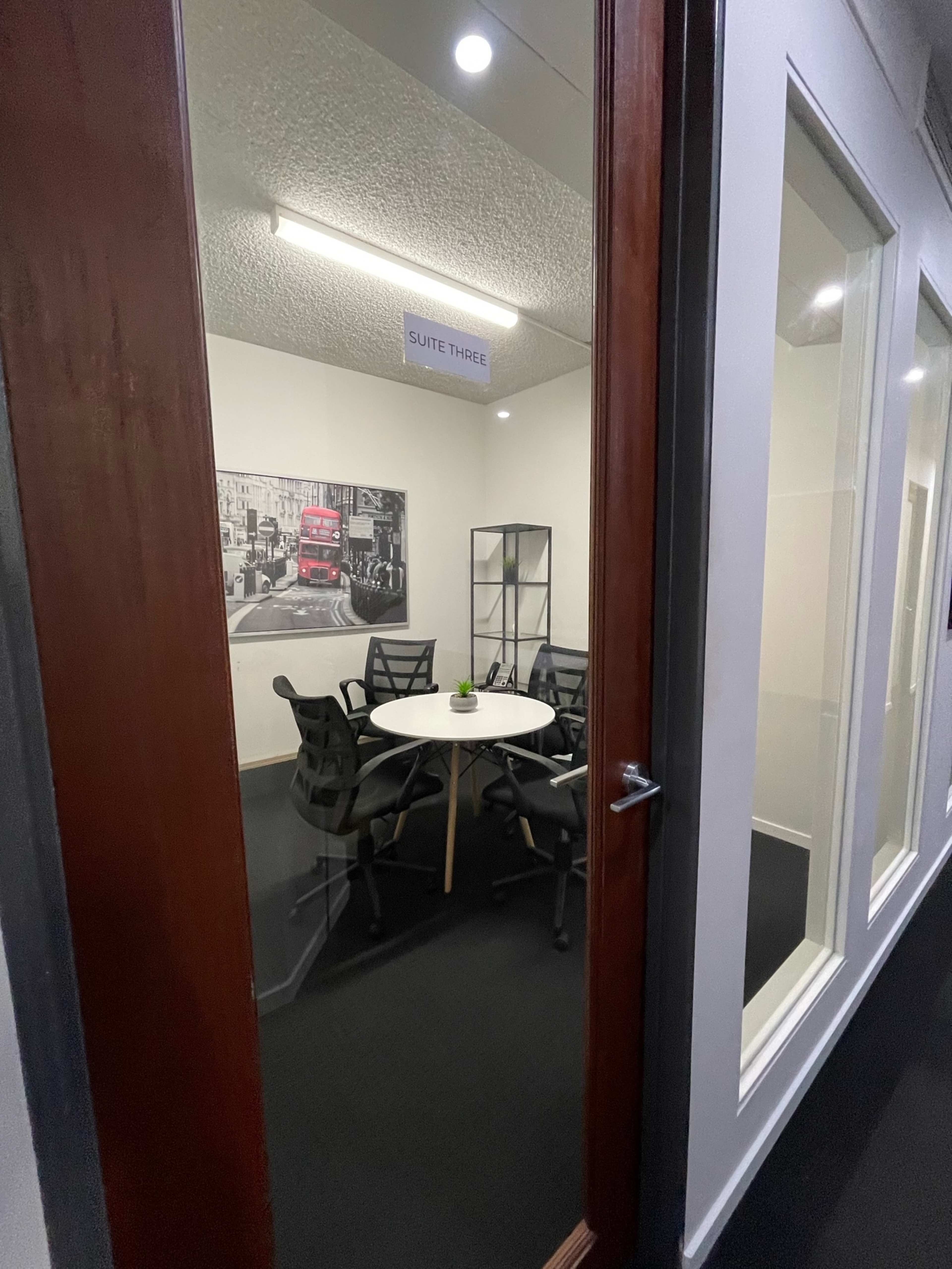 The image shows a modern office meeting room with a round table and black chairs, visible through a glass door labeled "Suite Three."