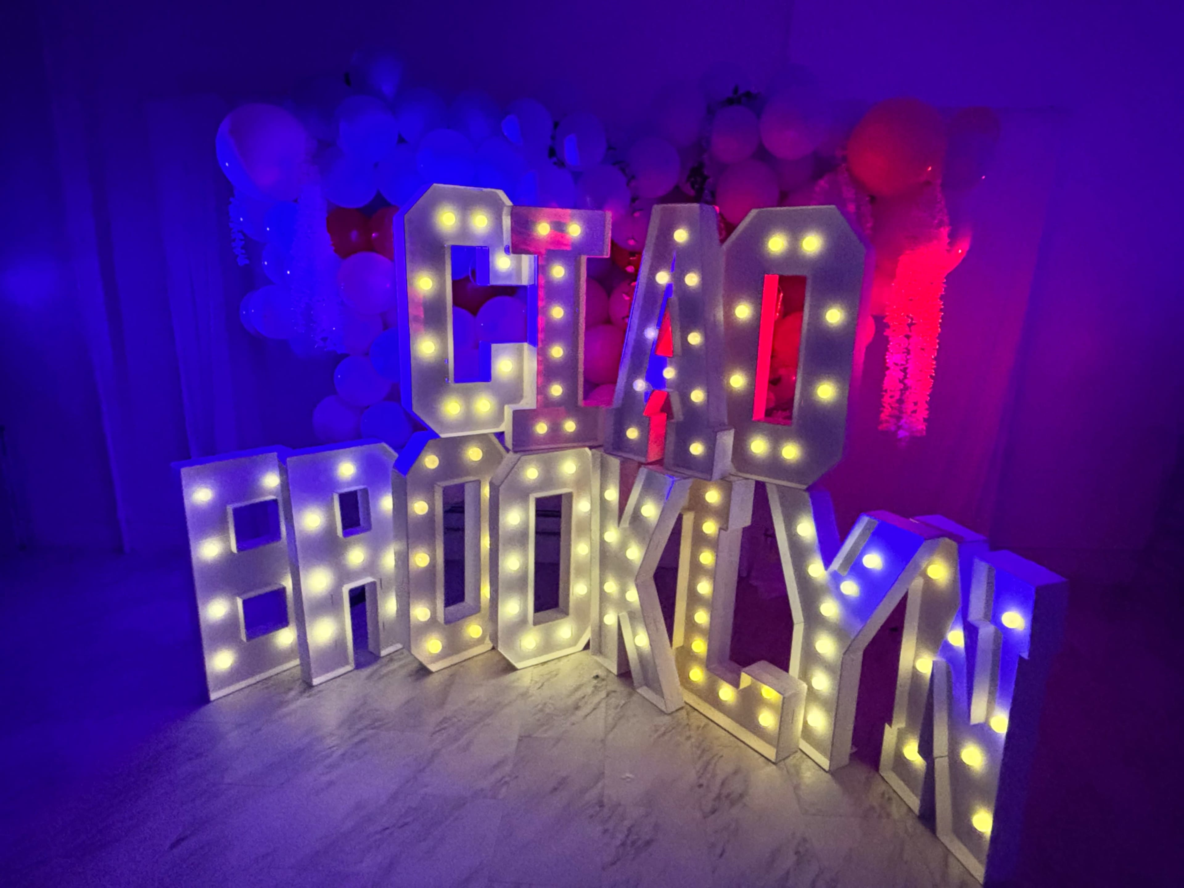 The image features a large illuminated sign that spells out "CIAO BROOKLYN," surrounded by colorful balloons and ambient lighting.