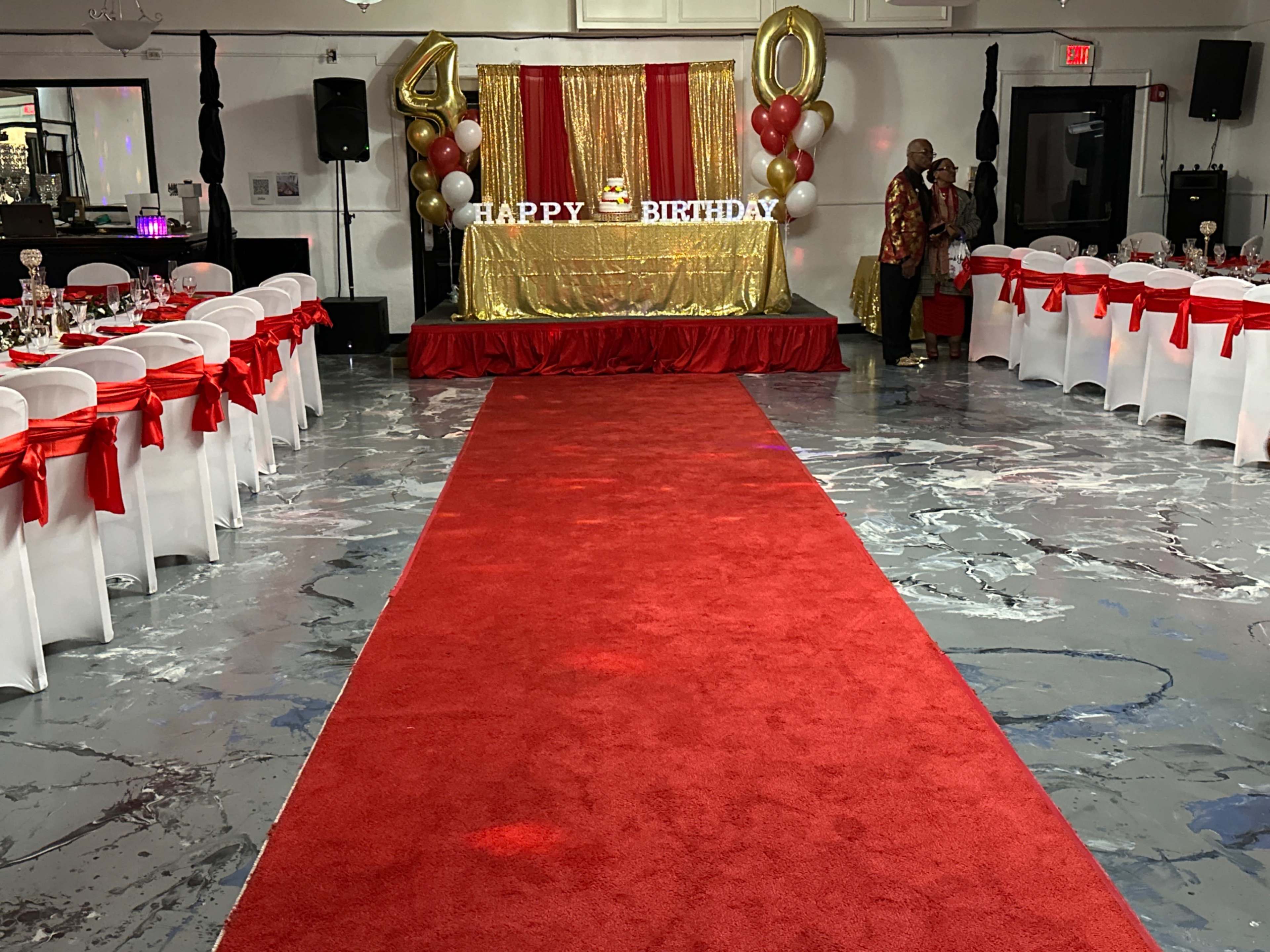 A red carpet leads to a birthday backdrop adorned with gold decorations and balloons for a 40th birthday celebration, with tables set on either side.