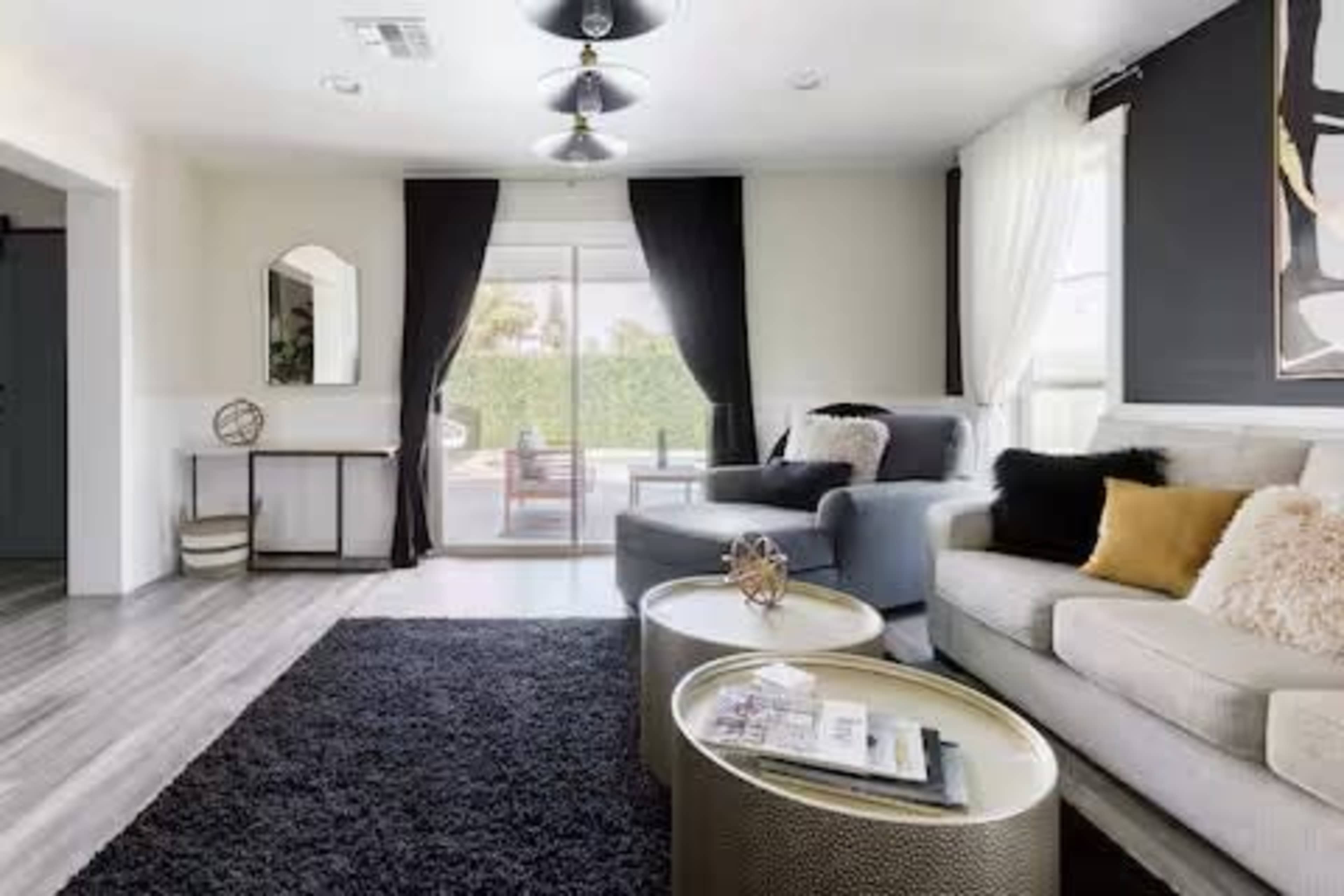 The image shows a modern living room featuring a gray sofa, a beige couch, two round coffee tables, and large windows with black curtains.