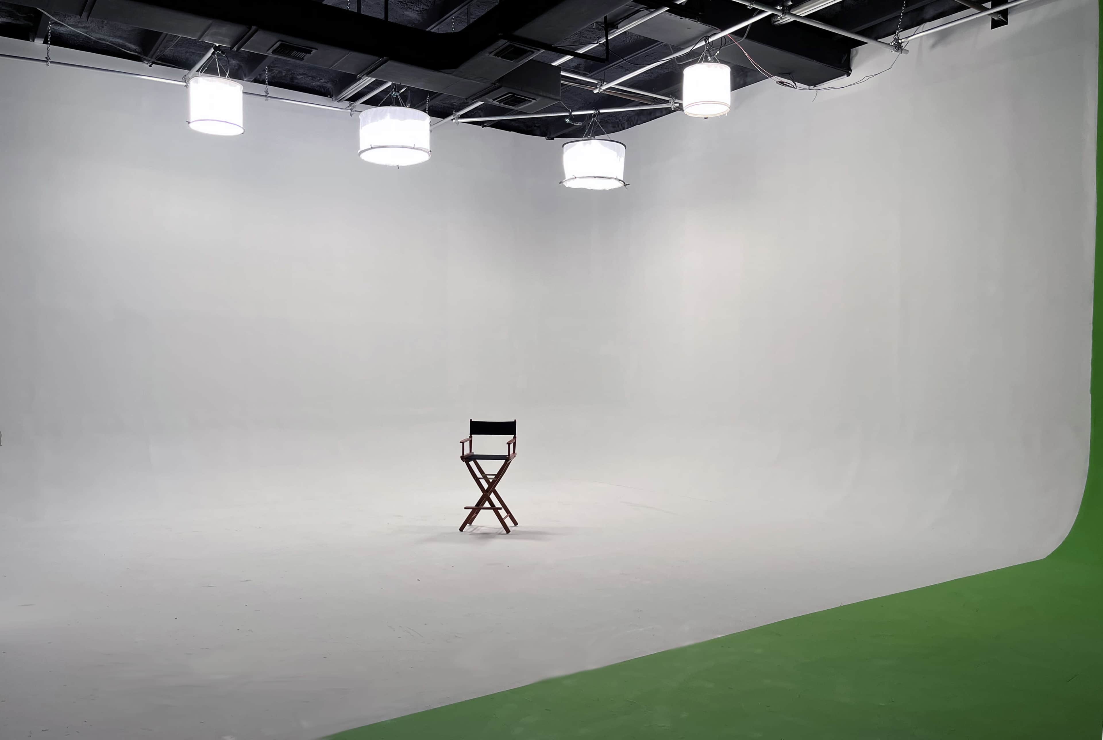 The image shows an empty film studio with a green backdrop, a white shooting area, and a single director's chair under overhead lights.