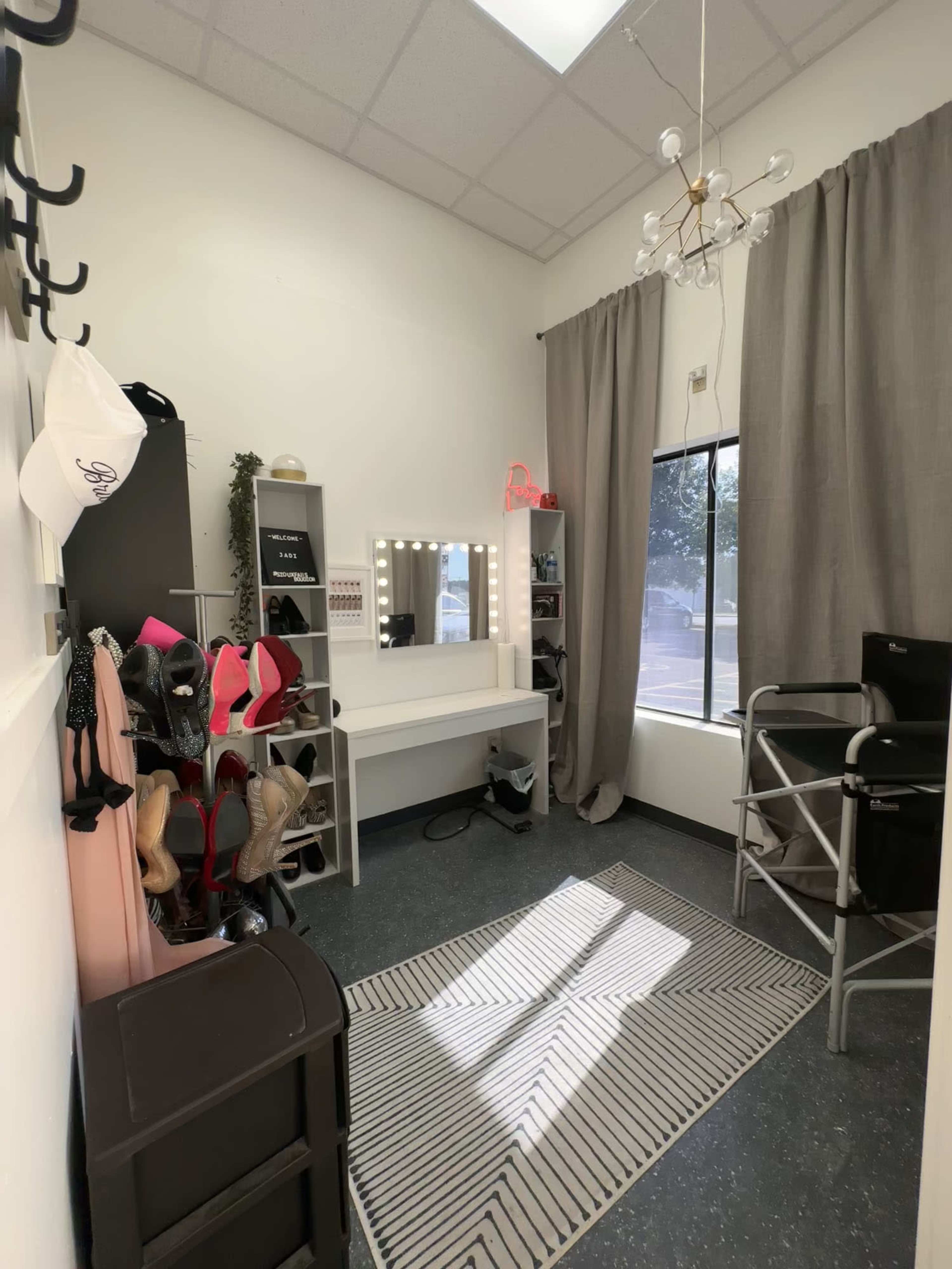 A well-organized room featuring a mirror with lights, a chair, a shoe rack filled with various shoes, and a window with curtains.