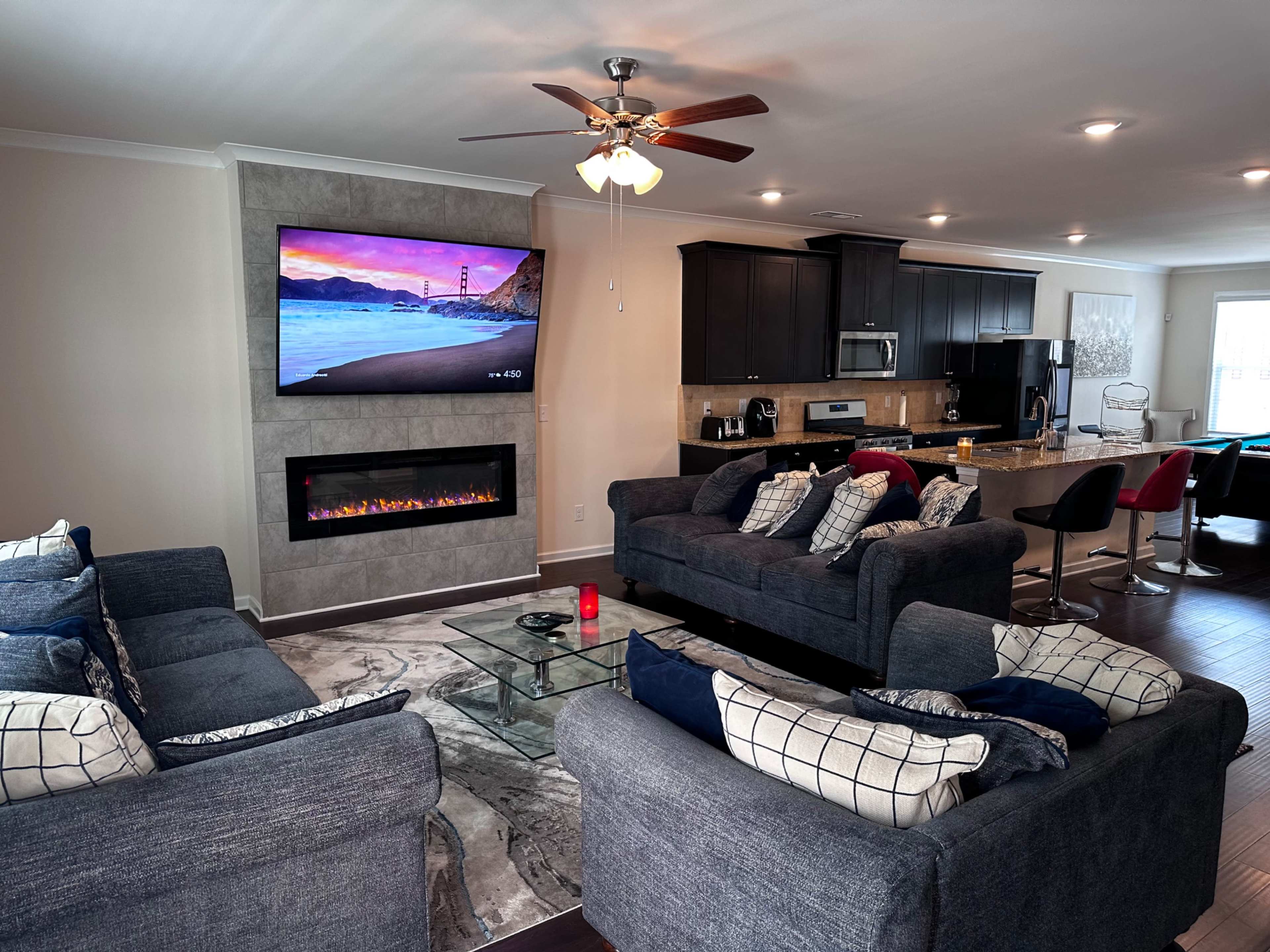 A modern living room featuring two gray sofas, a mounted television above an electric fireplace, and an open kitchen area in the background.