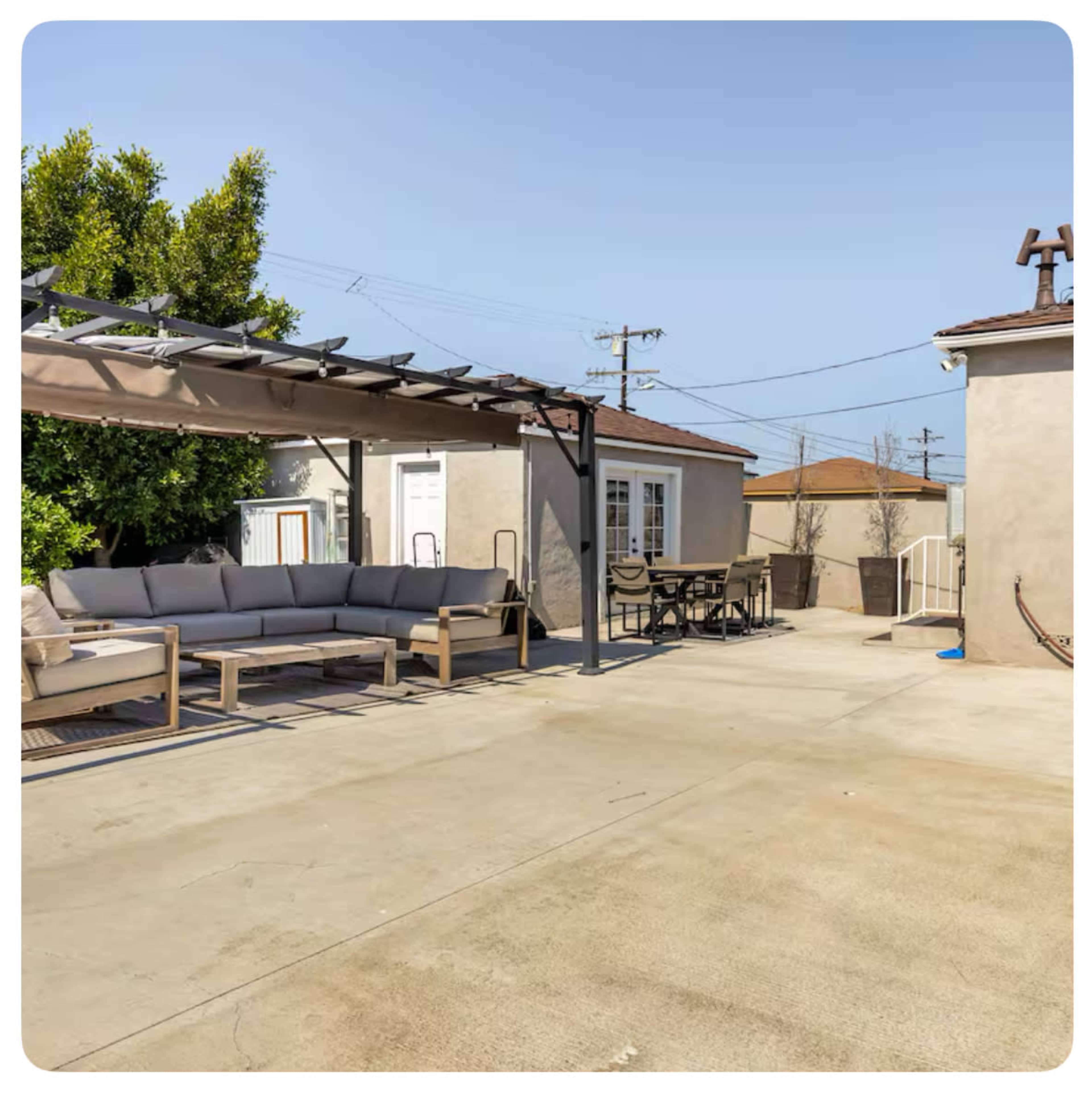 The image shows a patio area with a seating arrangement under a pergola and a table set nearby, surrounded by light-colored walls and utility poles in the background.