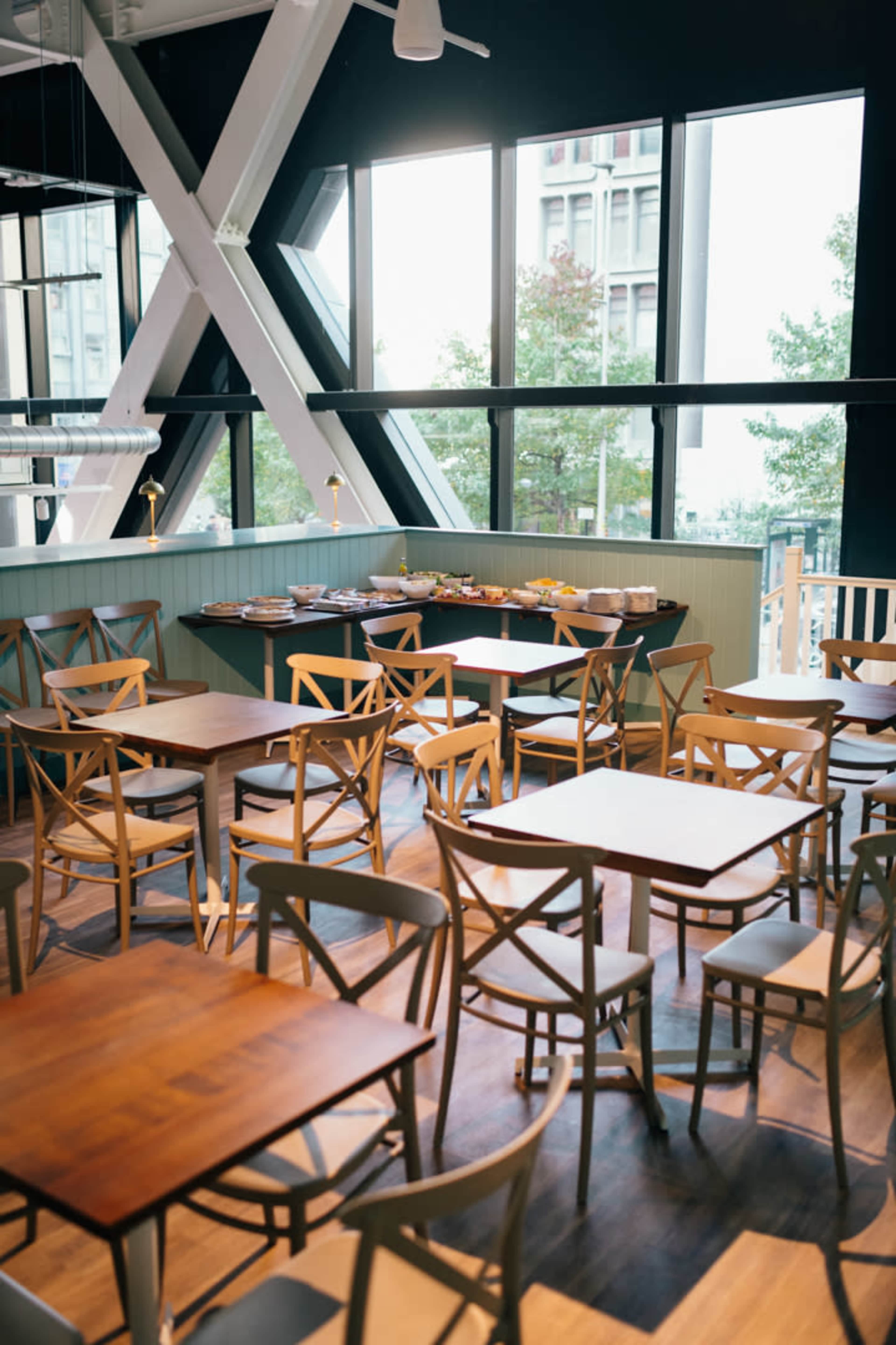 The image shows an empty restaurant interior featuring wooden tables and chairs, with large windows allowing natural light to fill the space.