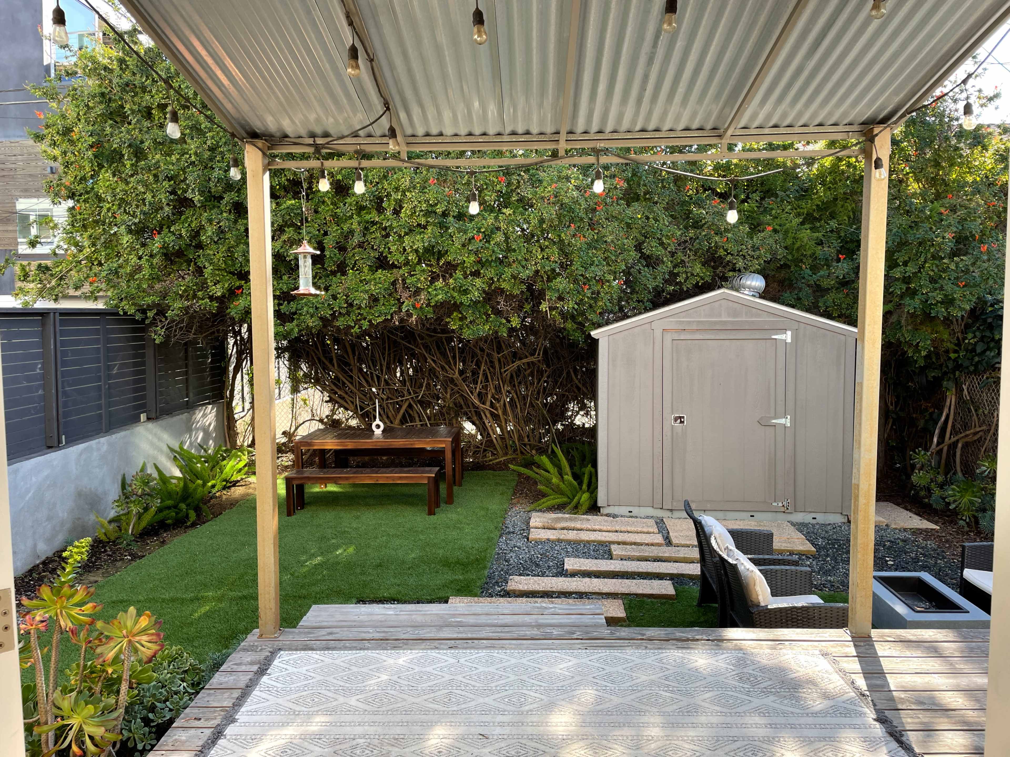 The image shows a backyard with a wooden table, a shed, a lush green lawn, and outdoor seating under a covered patio with decorative lights.