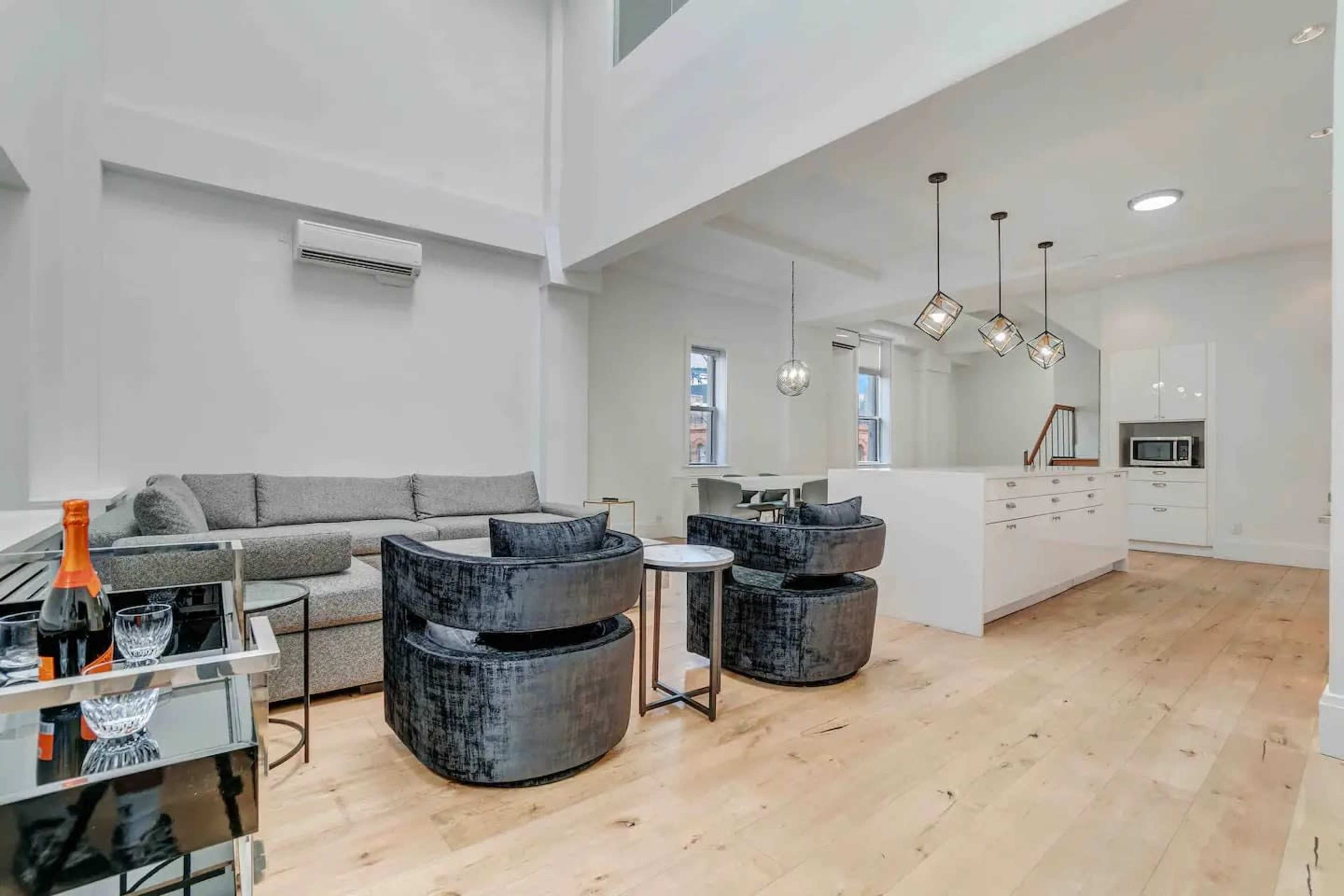 A modern living area with a gray sofa, two black chairs, a small table, and a kitchenette with white cabinetry and pendant lighting.