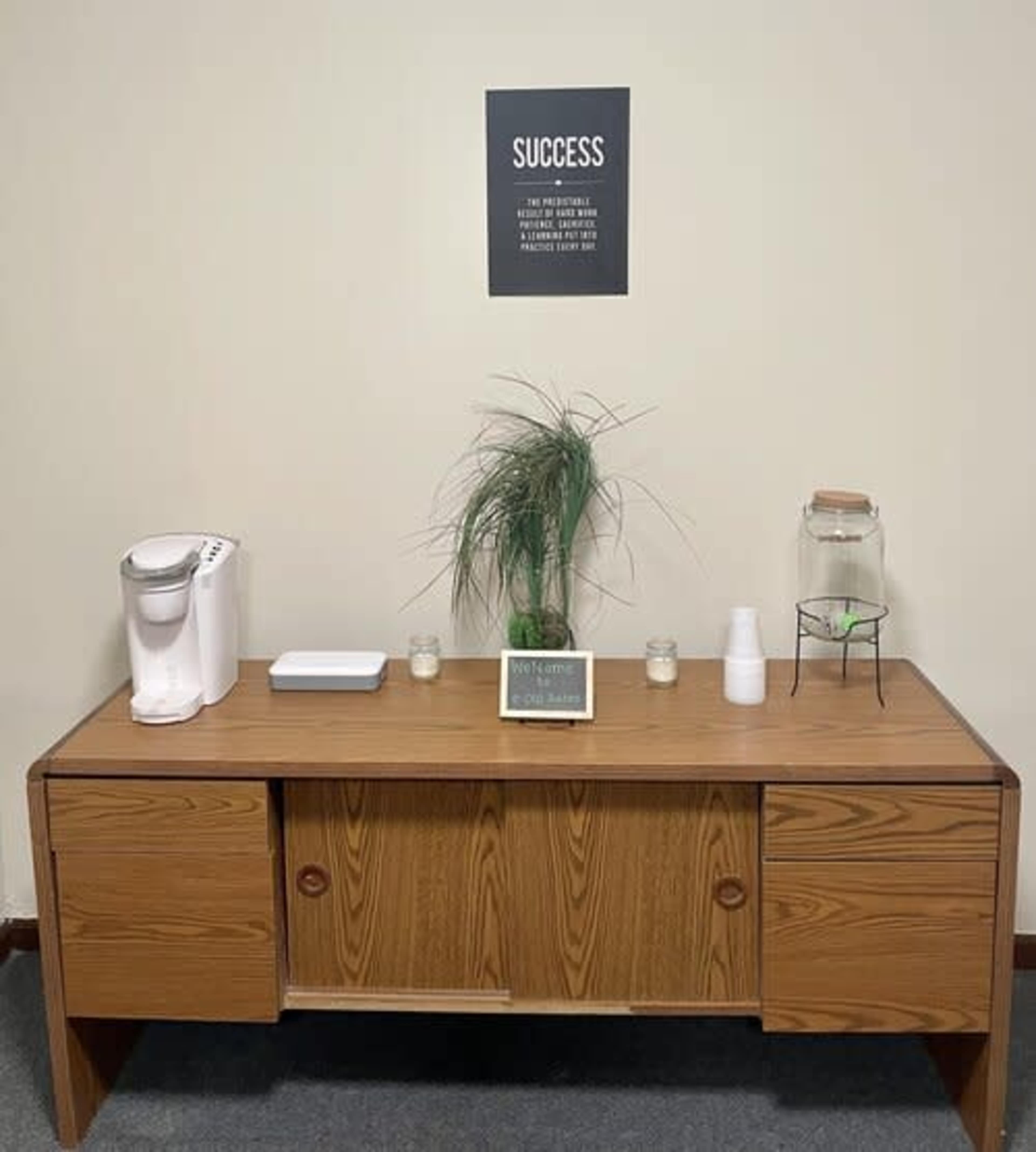 A wooden desk is displayed with a coffee maker, a plant, a small sign, and candles against a simple wall featuring a motivational poster that reads "SUCCESS."