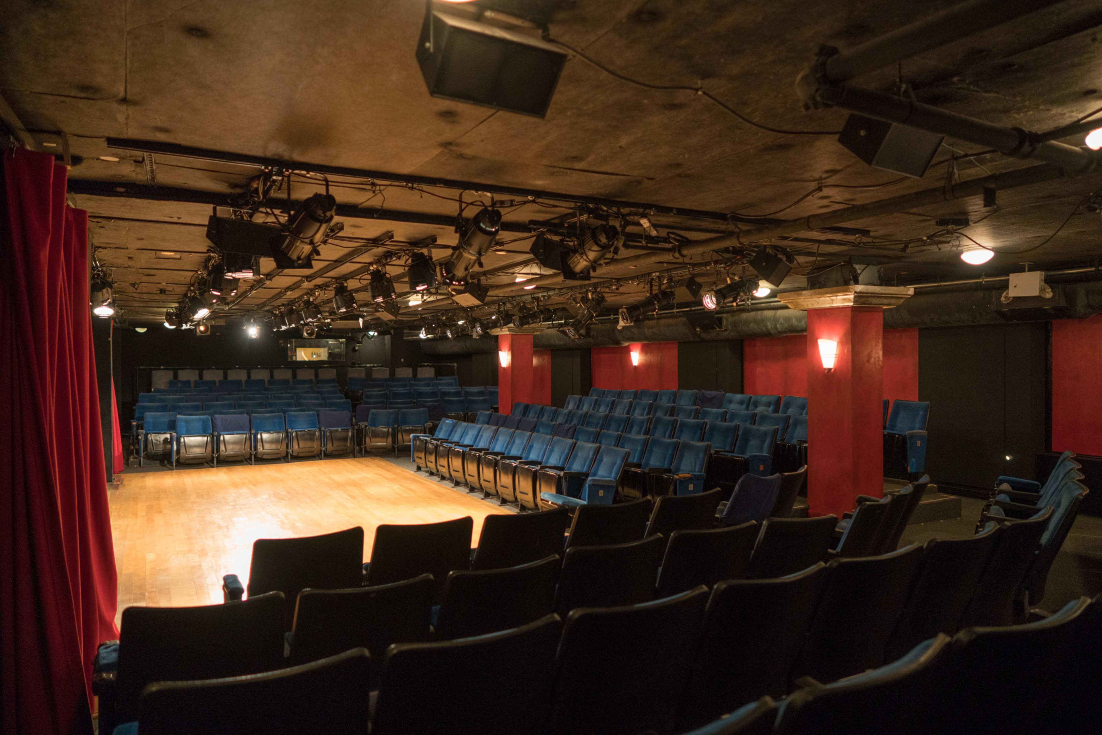The image shows a small theater with blue seating arranged in rows facing a wooden stage, illuminated by overhead lights.