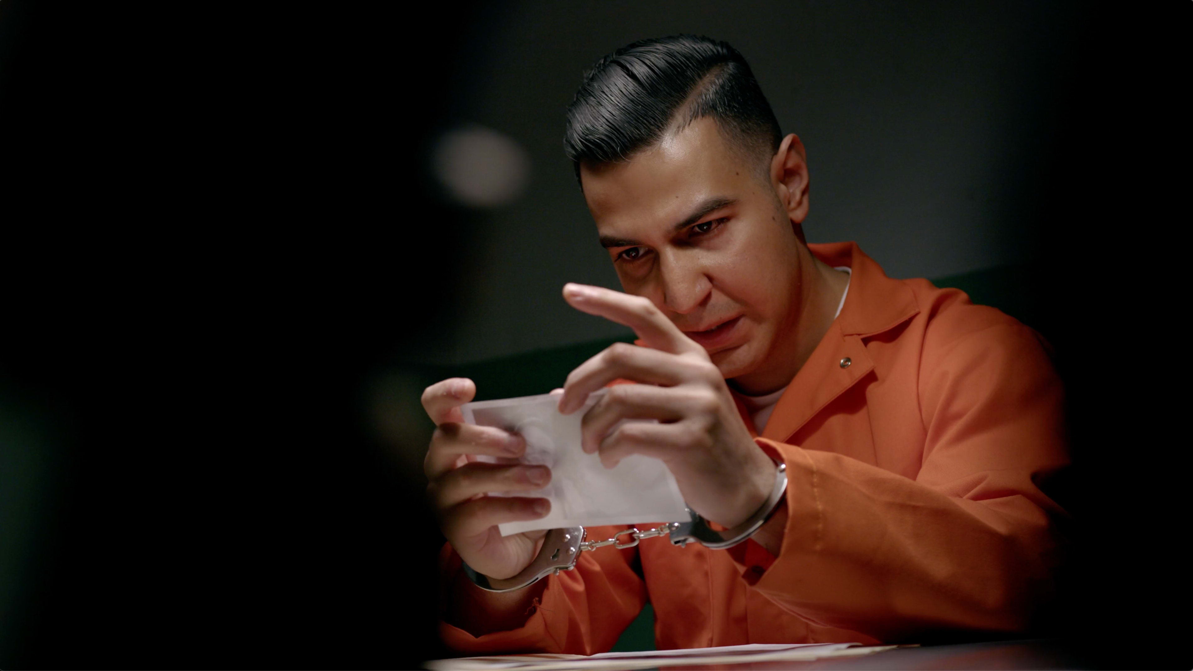 A man in an orange prison jumpsuit examines a piece of paper while seated at a table with his hands in handcuffs.