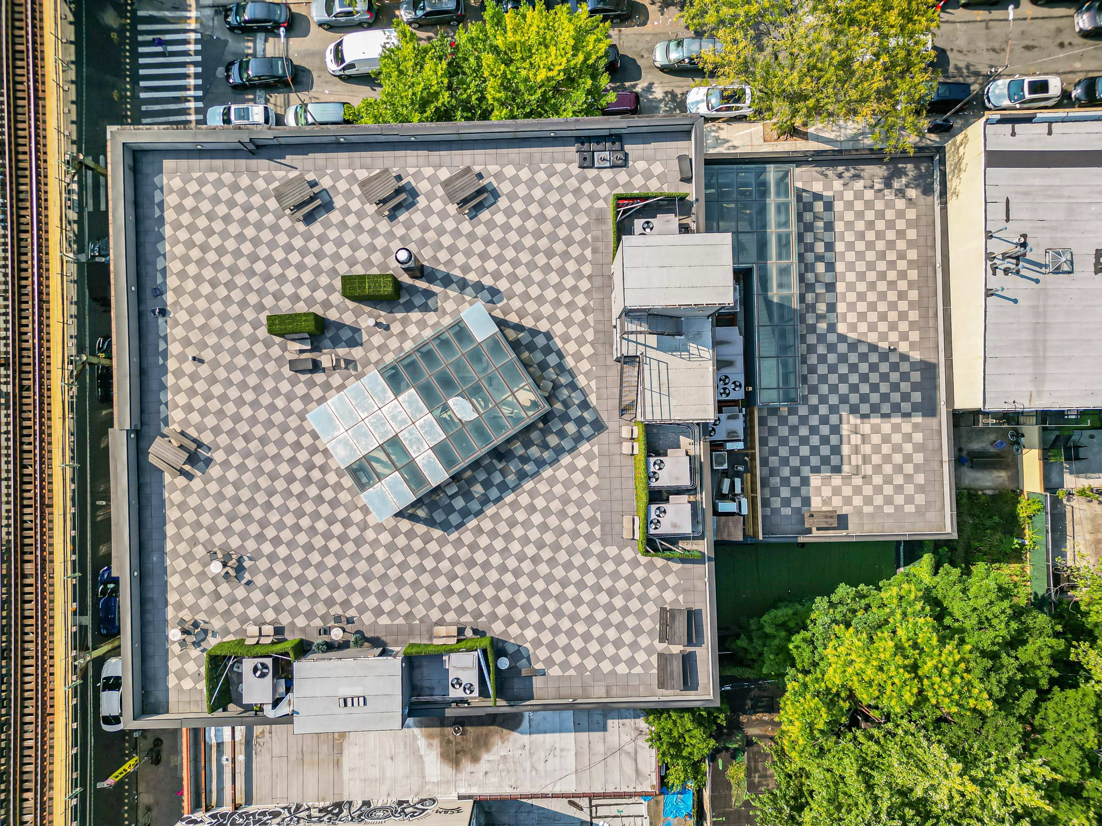An aerial view of a building rooftop with a checkerboard pattern, featuring seating areas and vegetation, adjacent to parked cars and a railway track.