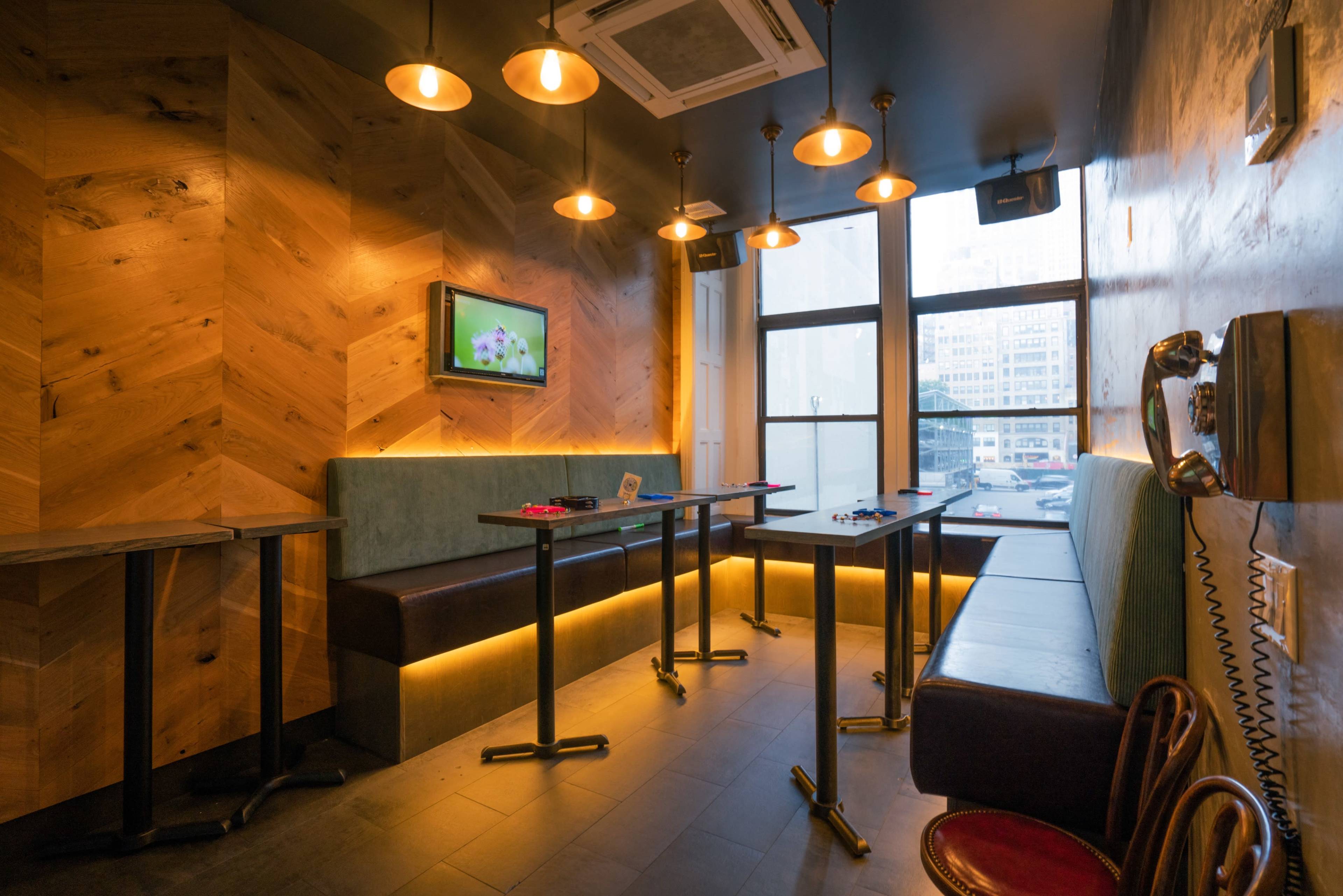 A cozy interior features wooden walls, a small television, hanging lights, and tables along the windows with a city view.