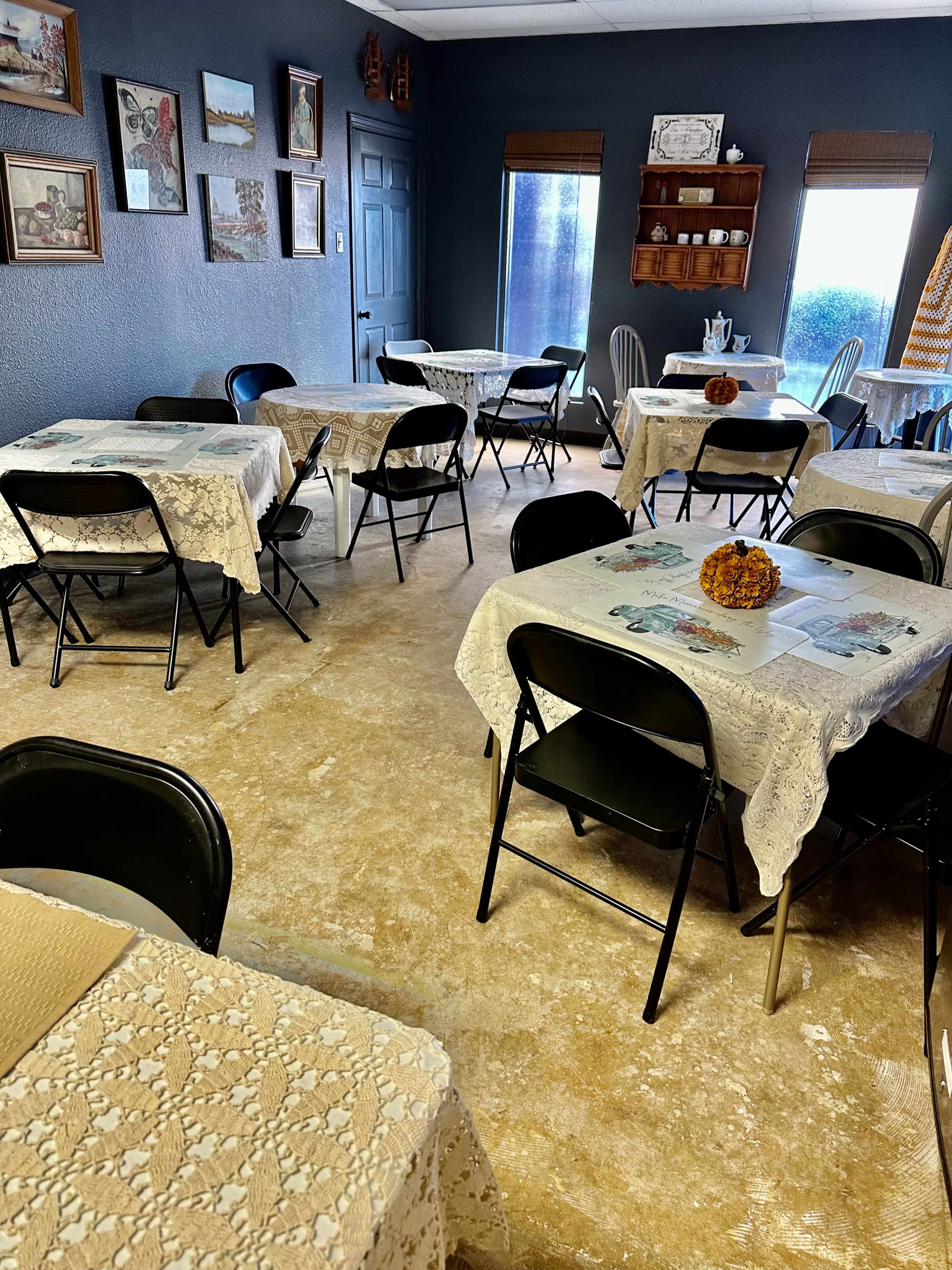 A dining area with several tables covered in lace tablecloths, surrounded by black folding chairs and decorated with wall art.