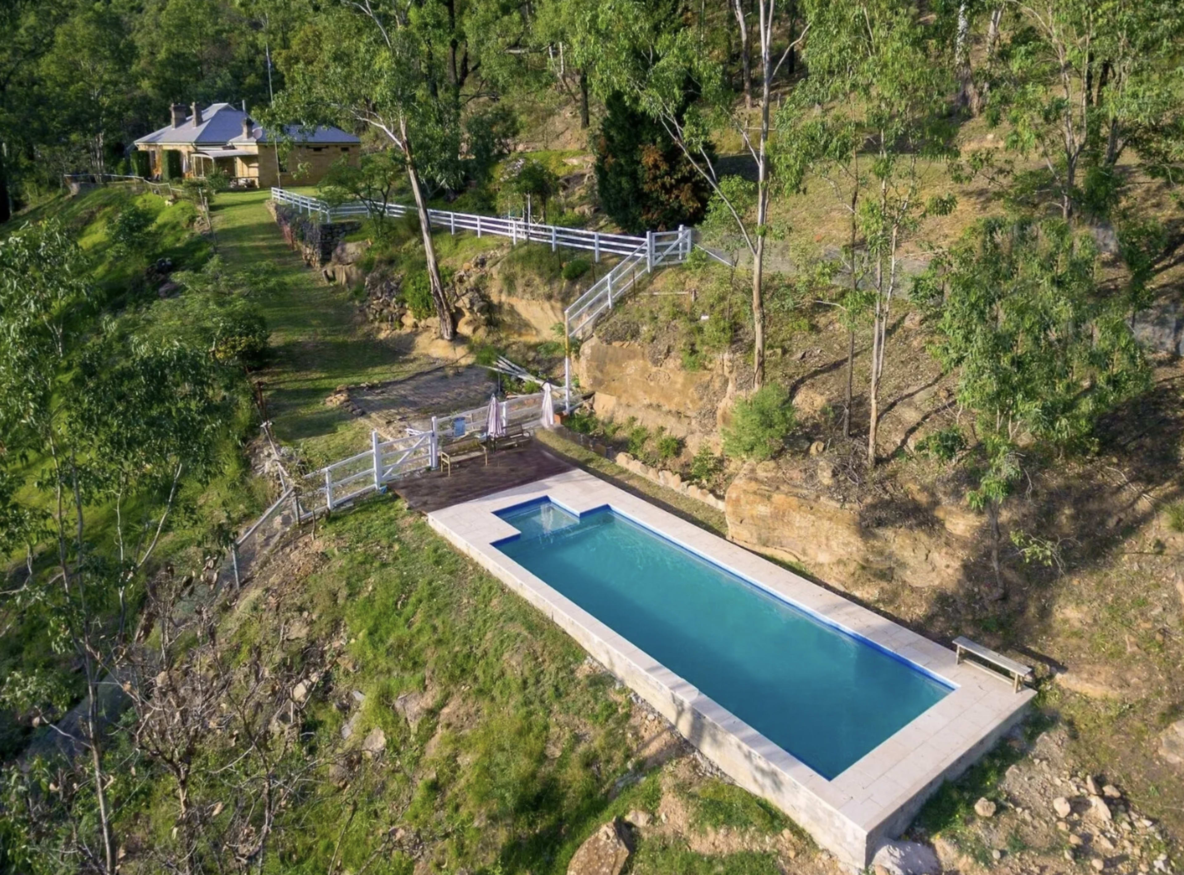 A rectangular swimming pool situated on a hillside, with a wooden deck and a house visible in the background, surrounded by greenery.