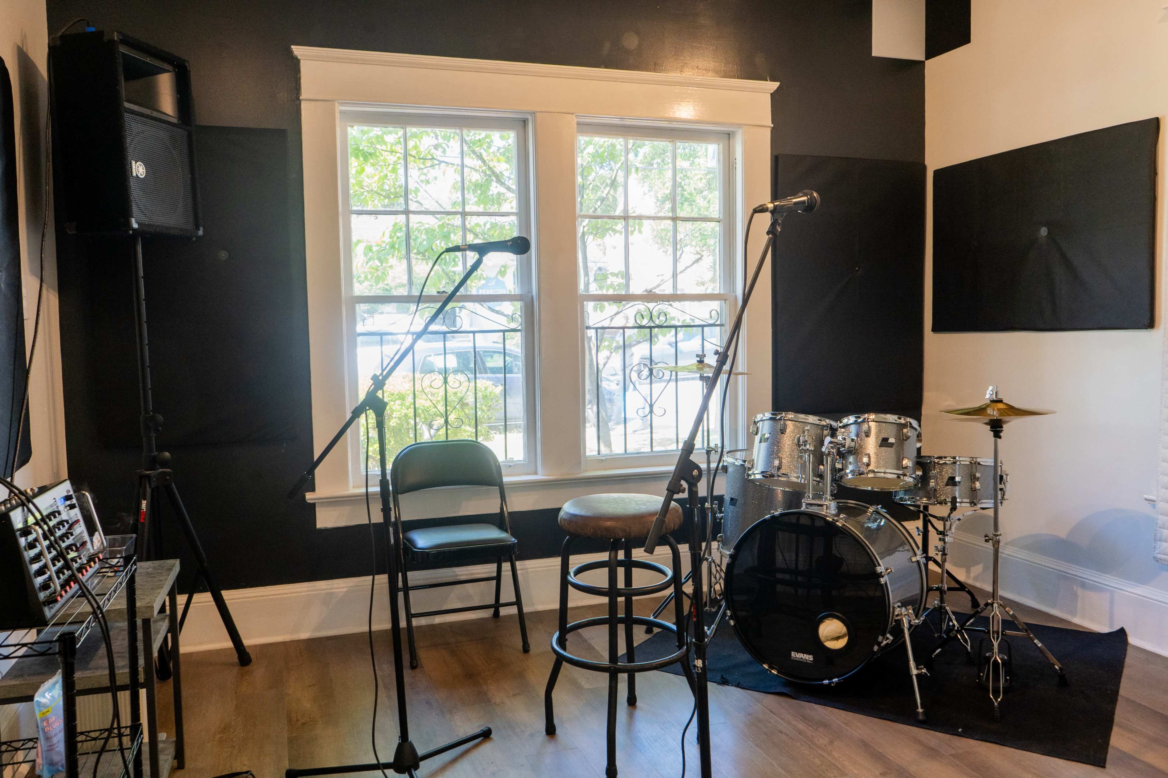 A small practice room is equipped with a drum set, two microphones on stands, and a chair near a large window.
