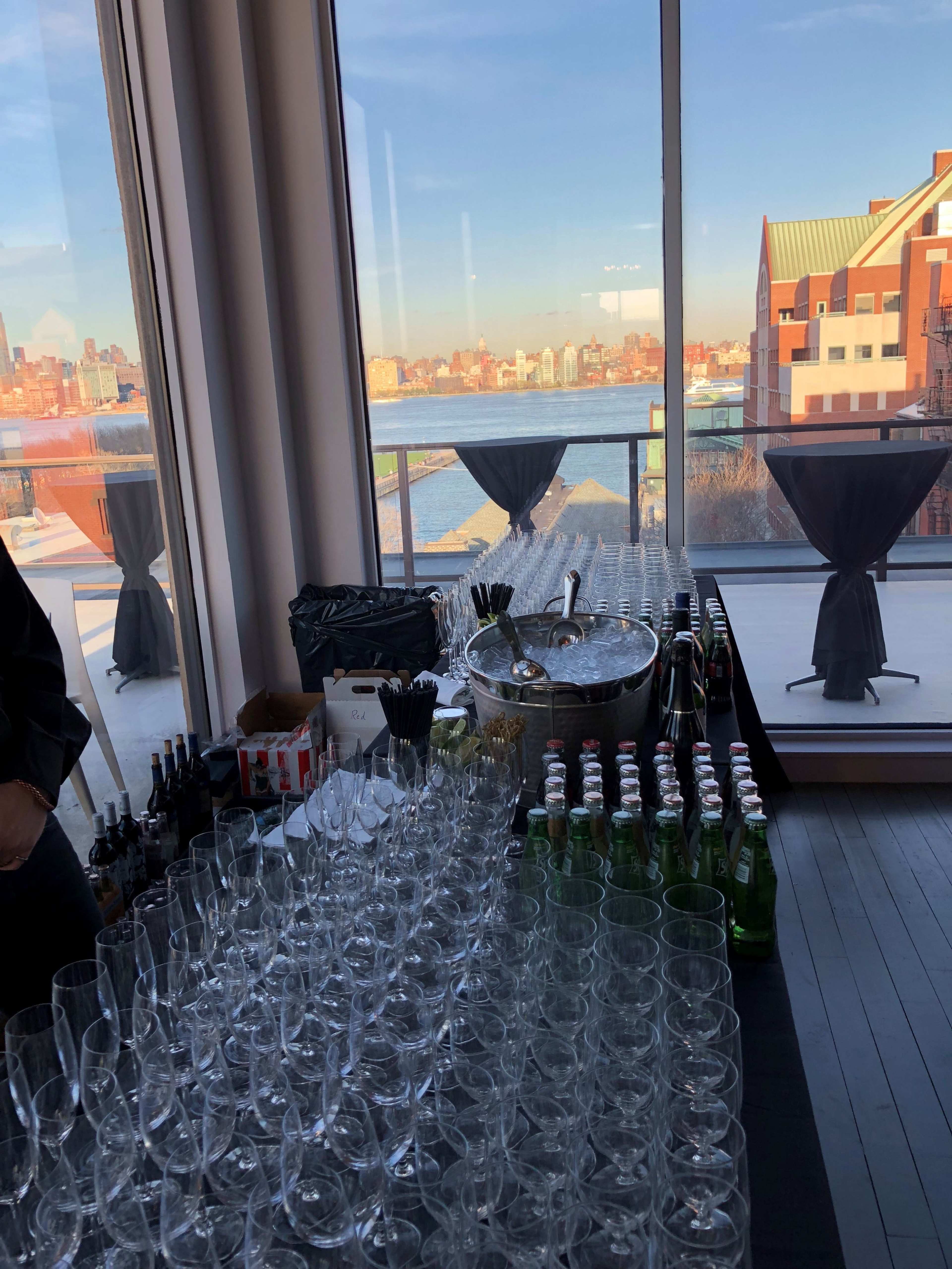 A table is filled with neatly arranged glassware and bottles, set against a backdrop of a waterfront view and city skyline.