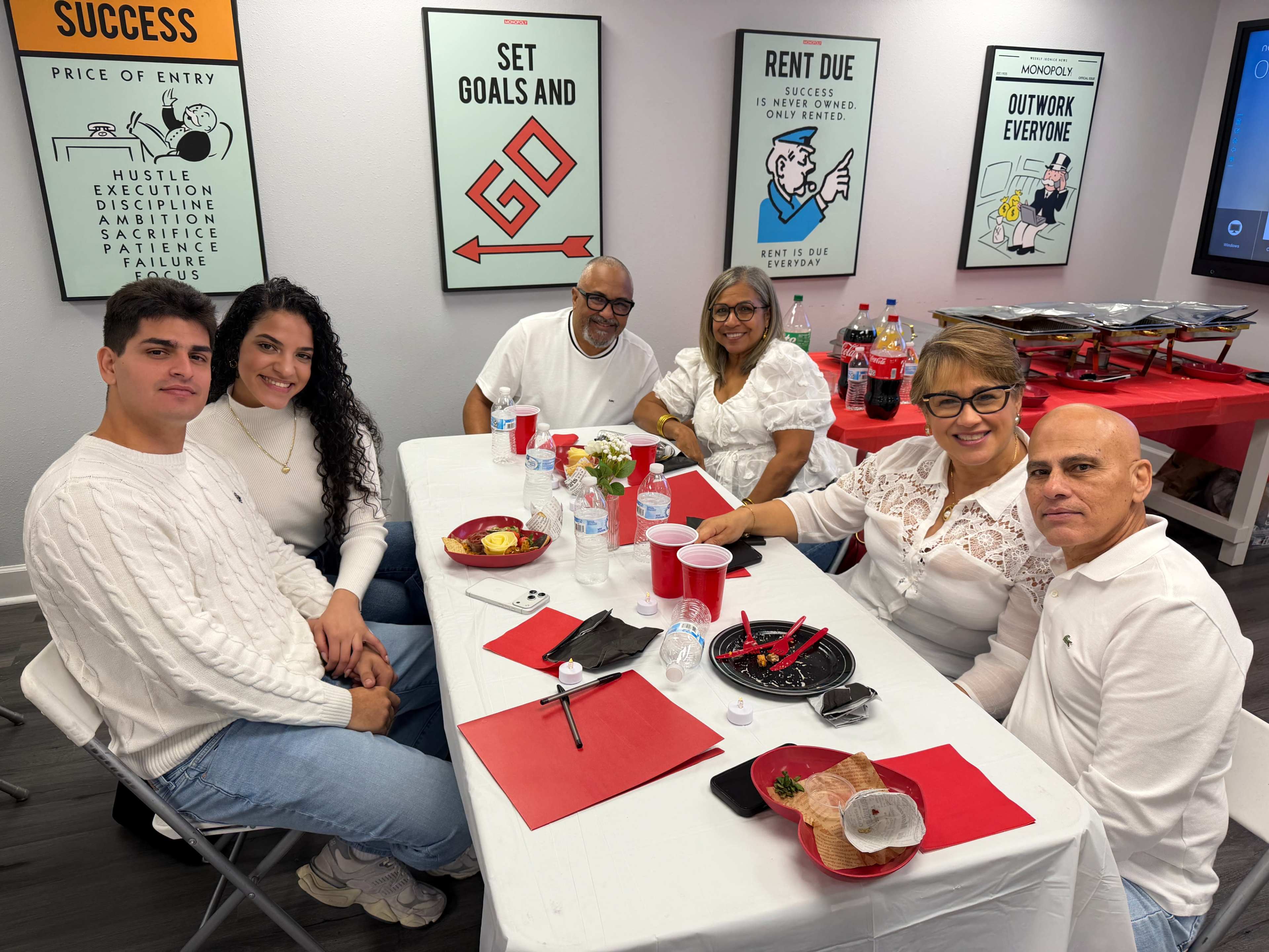 A group of six people sits at a table decorated with red and white items, surrounded by motivational posters and a spread of food in a room.