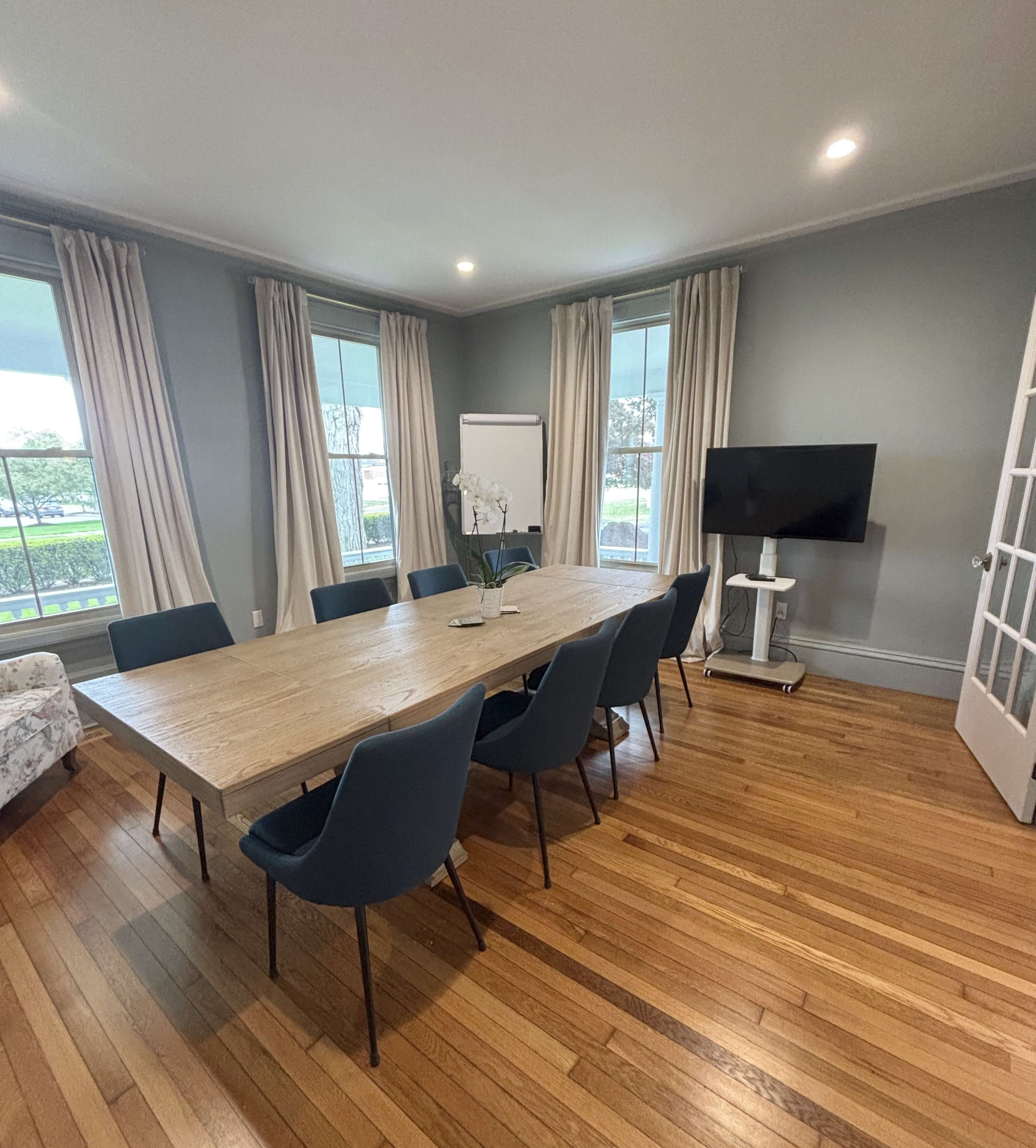 The image shows a conference room with a long wooden table surrounded by blue chairs, a television, and large windows with curtains.
