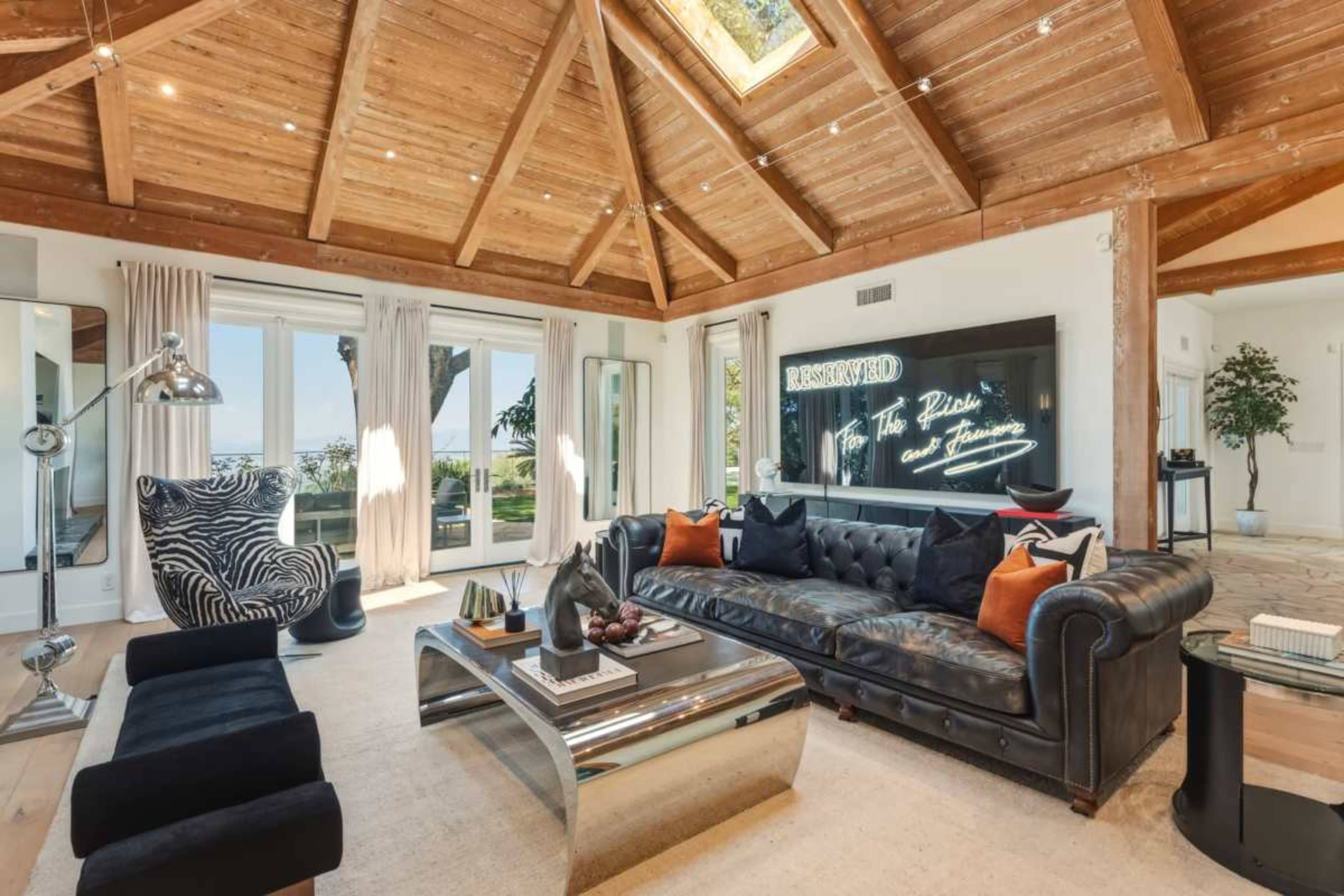 The living room features a high, wooden-beamed ceiling, a large black leather sofa, a zebra-patterned chair, and expansive windows offering a view of the outdoors.