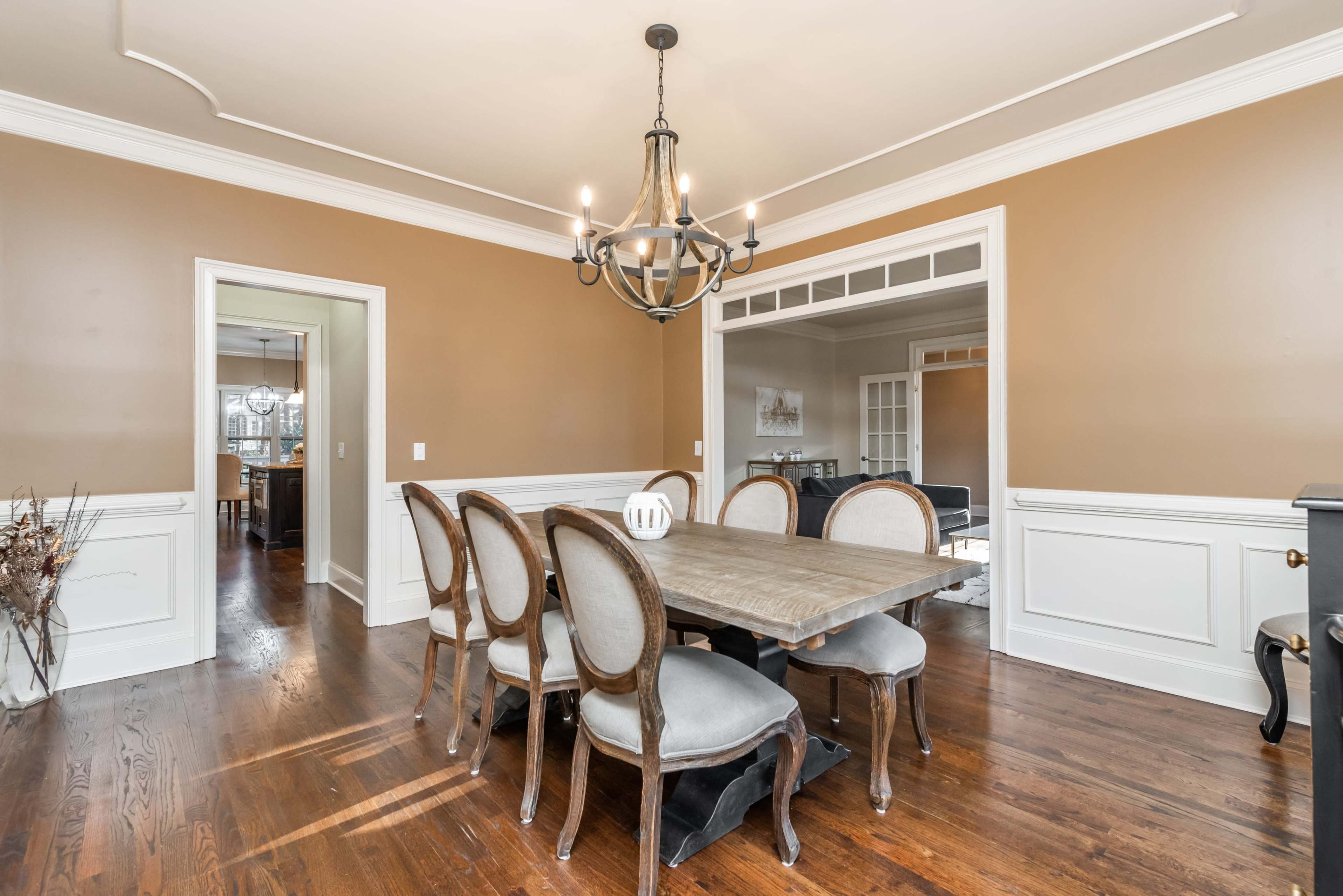 A dining room features a wooden table surrounded by six upholstered chairs, with a chandelier hanging above and warm-colored walls.