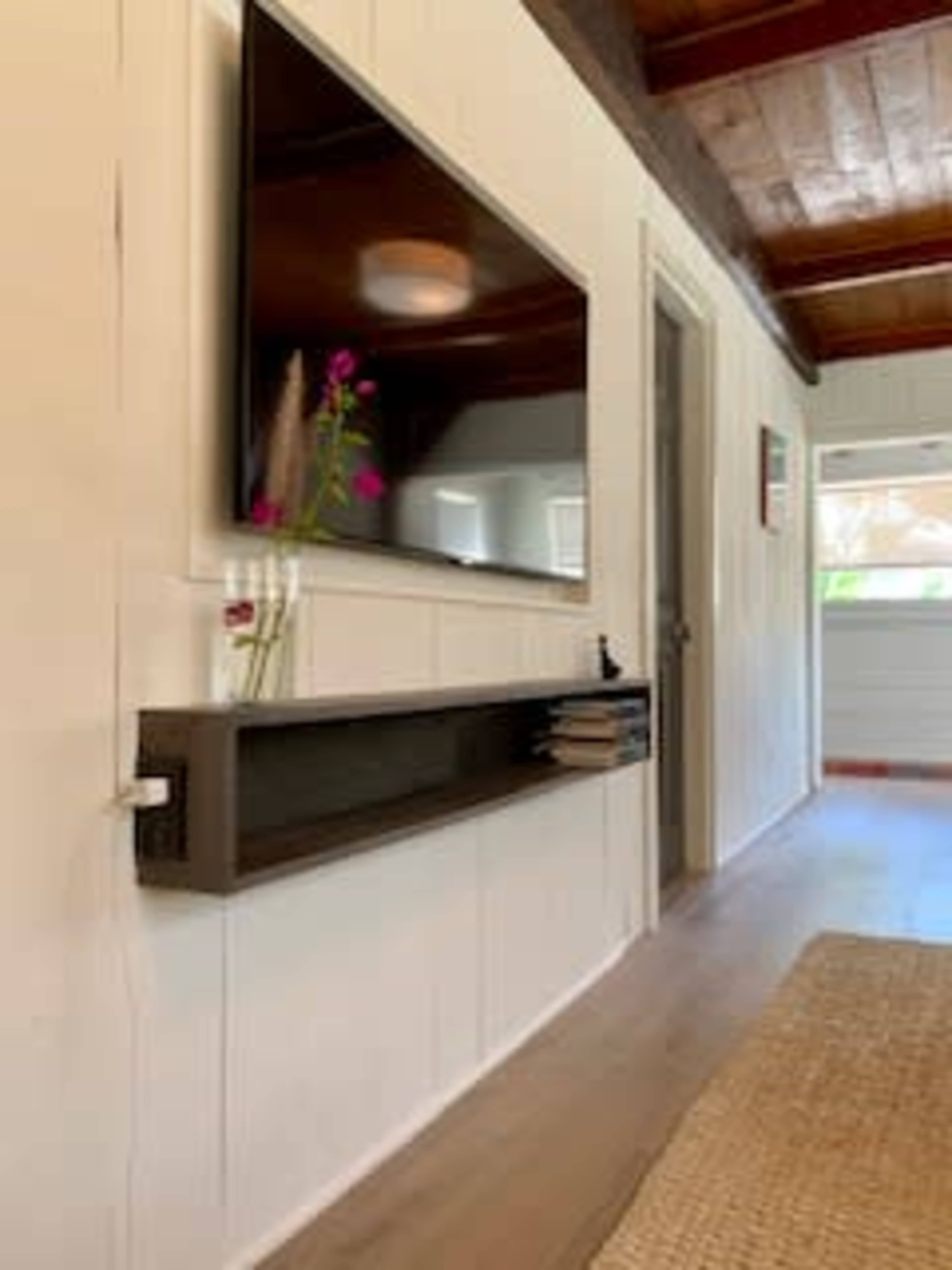 A flat-screen television is mounted on a white wall above a slim shelf holding a few books and a vase with flowers.