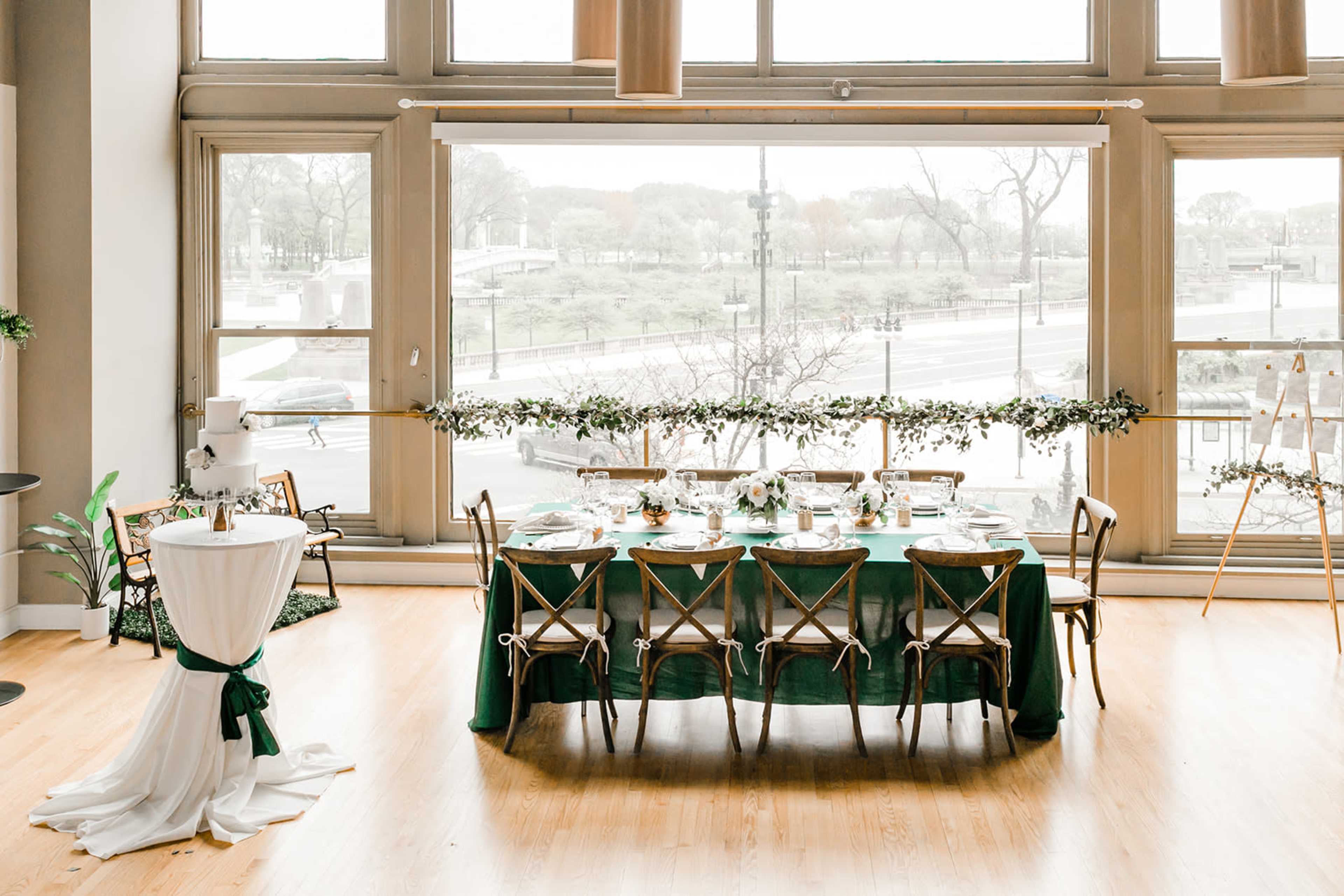 A long dining table is set with elegant tableware and greenery, positioned near large windows overlooking a park.