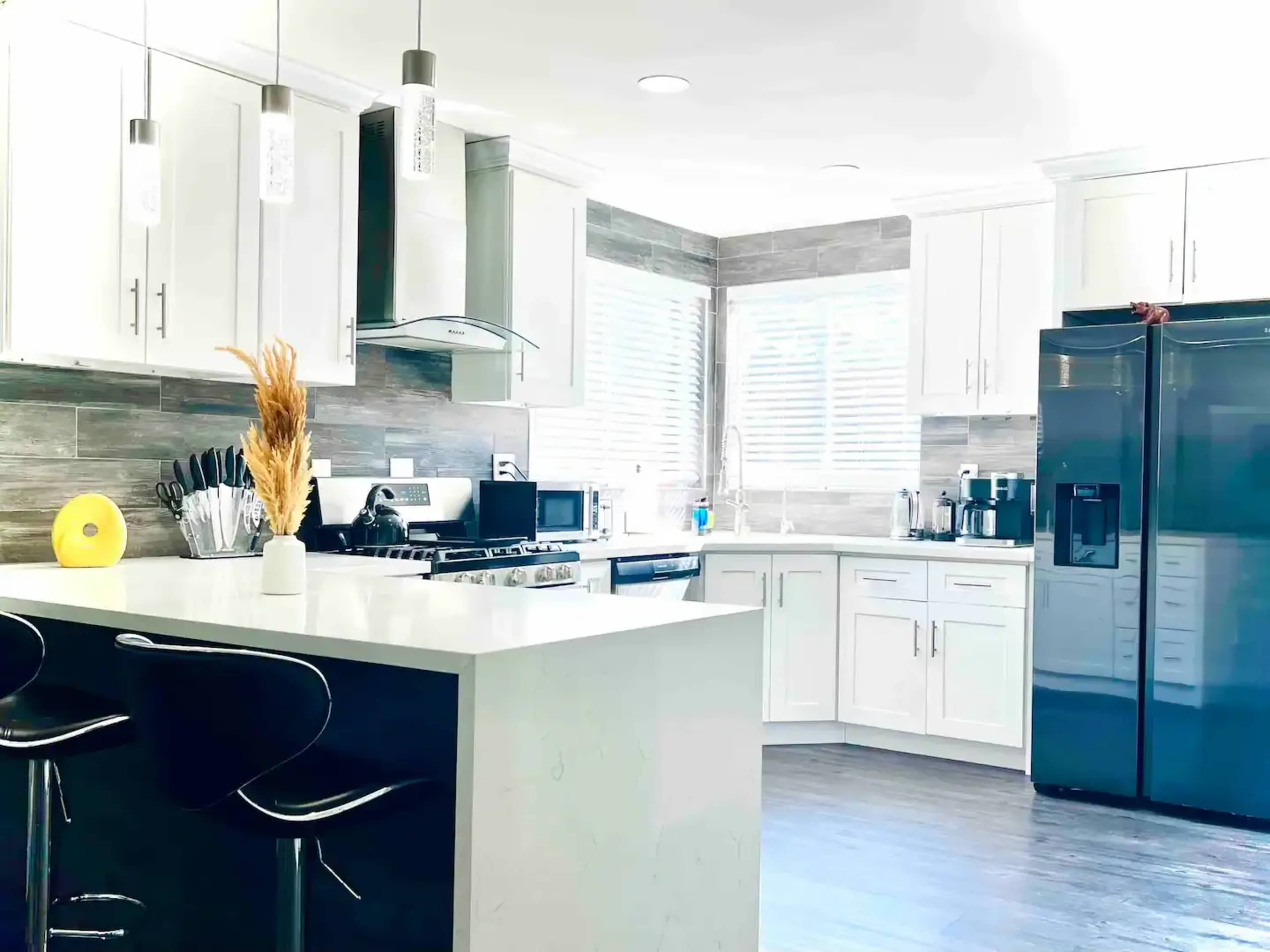 A modern kitchen features white cabinetry, stainless steel appliances, a large island with bar stools, and a dark wooden floor.