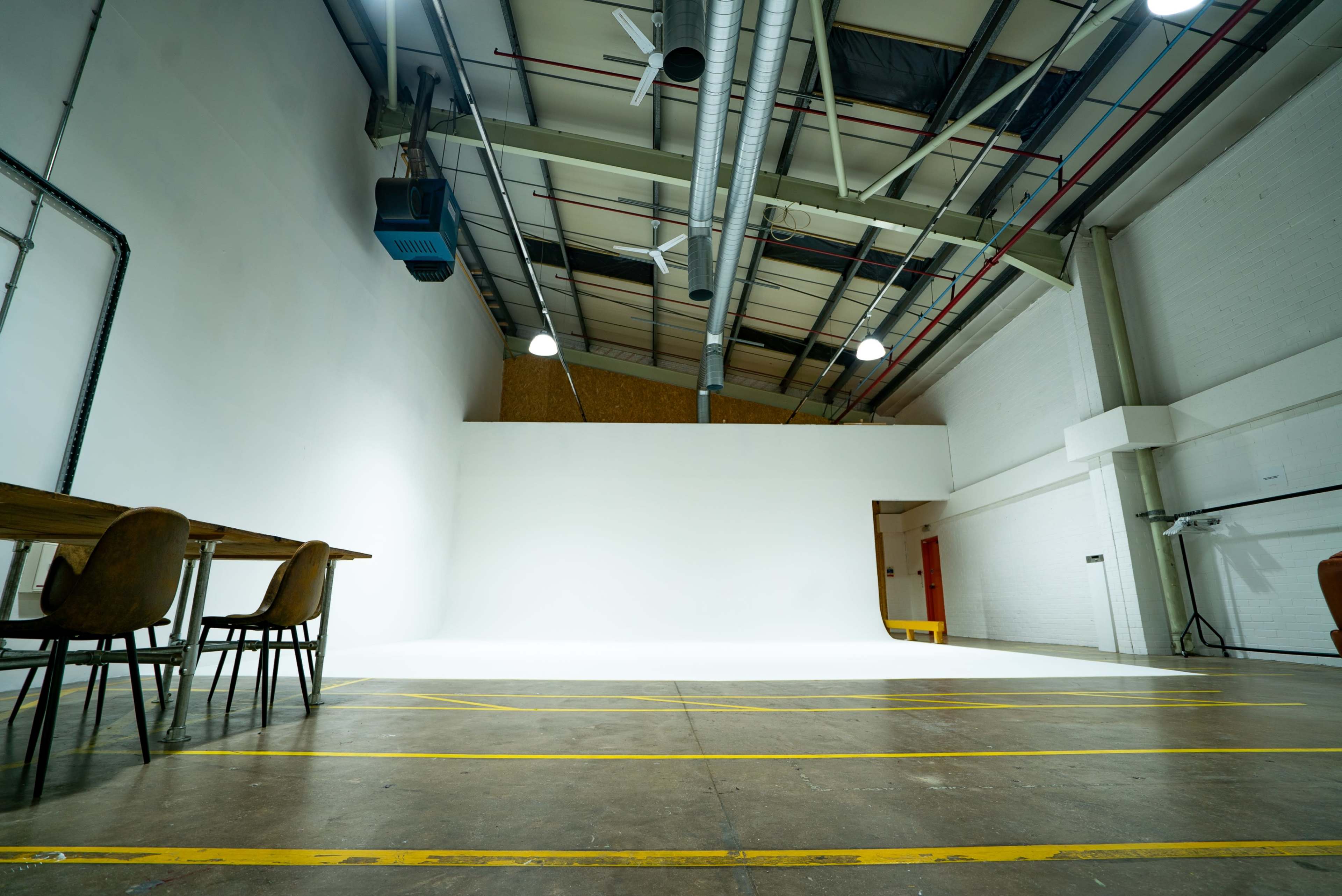The image shows a spacious studio with a clean white backdrop, wooden tables, and chairs, featuring exposed ceiling infrastructure and bright overhead lights.