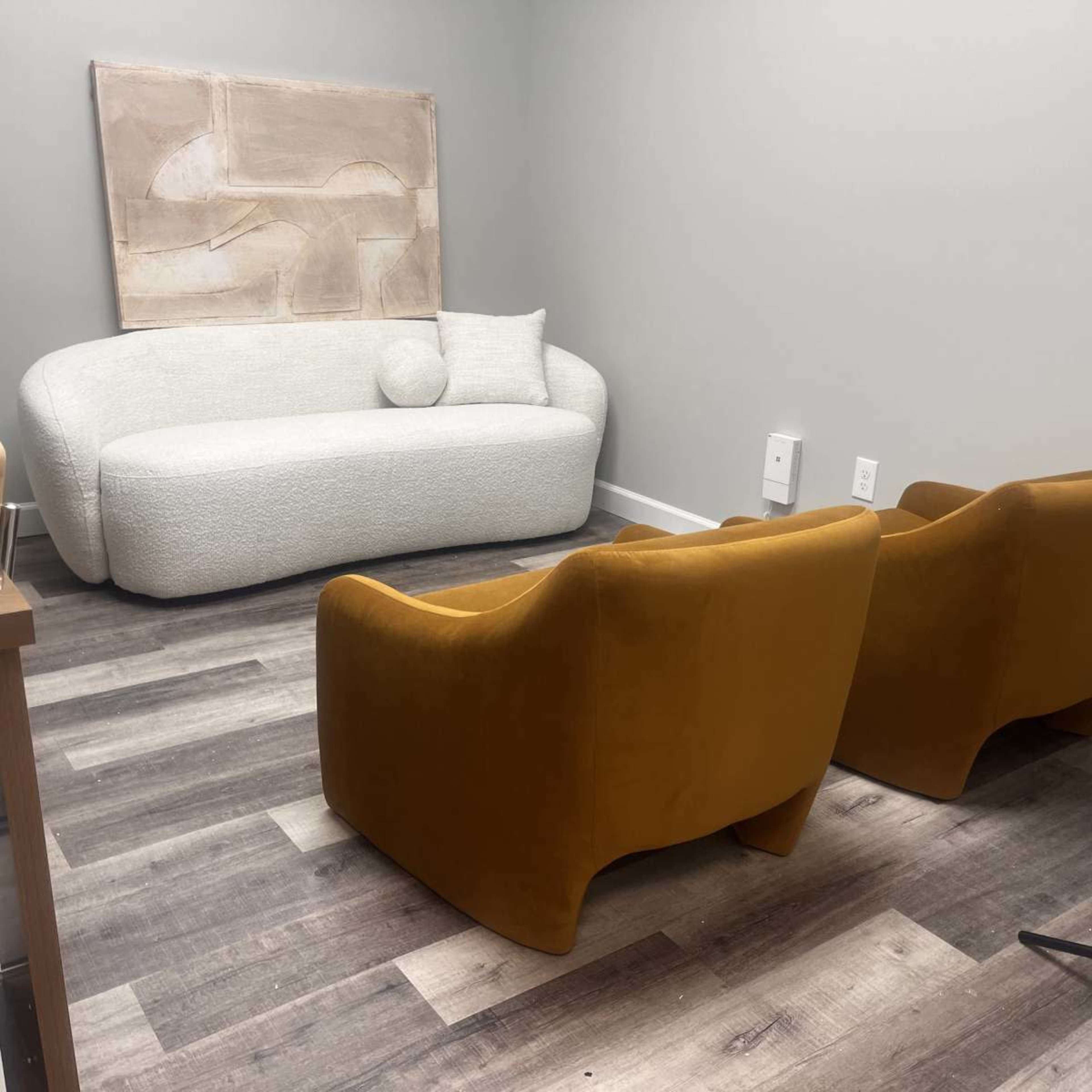 The image shows a minimalist room with a white, curved sofa, two yellow chairs, and an abstract artwork on the wall.