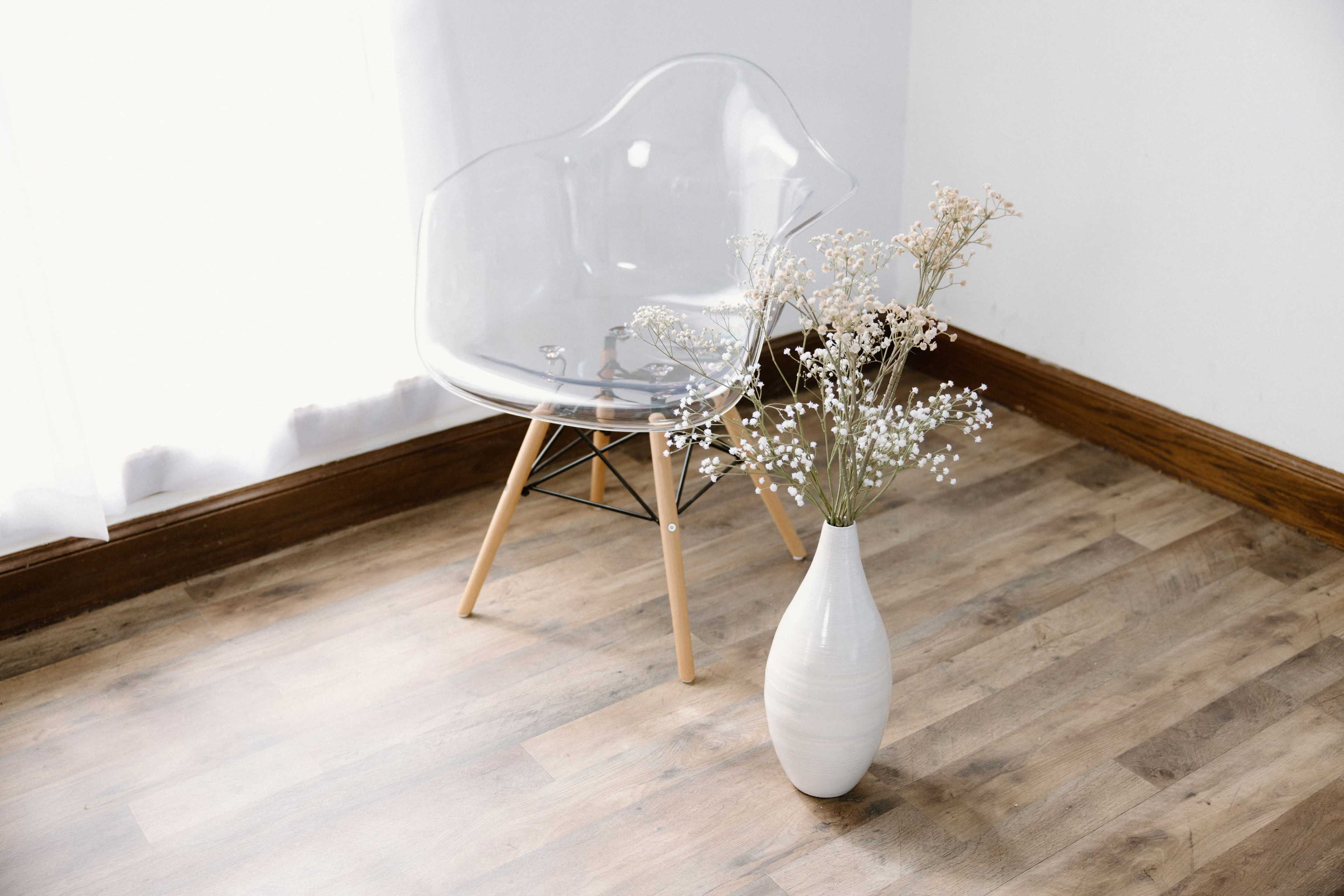 A clear plastic chair sits beside a white vase filled with dried flowers on a wooden floor near a window.